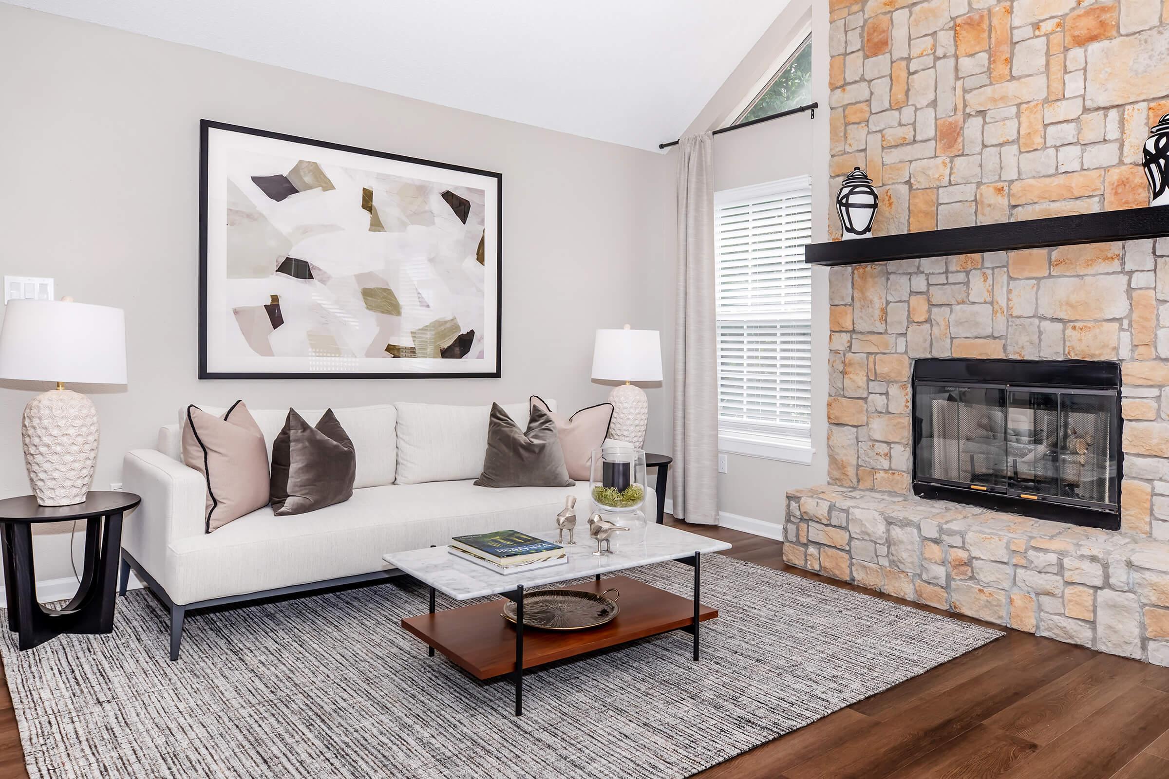 A modern living room featuring a light-colored sofa with decorative pillows, a marble coffee table, and two textured lamps on side tables. A stone fireplace is set into the wall, and a large abstract art piece hangs above the sofa, with neutral tones complementing the overall decor. Natural light fills the space through a window.