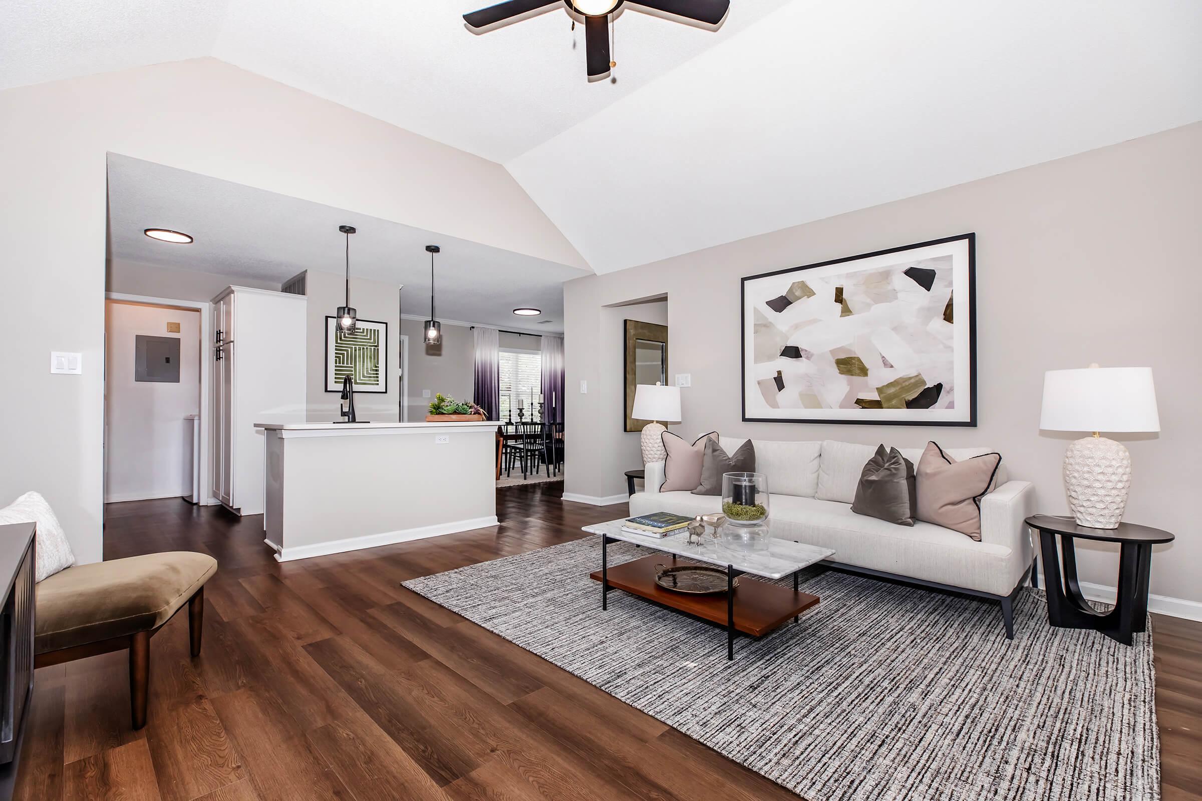 A modern living room featuring a light-colored sofa with decorative cushions, a coffee table, and a framed abstract artwork on the wall. The space has a stylish area rug and a ceiling fan, with soft lighting and a view into a dining area in the background.