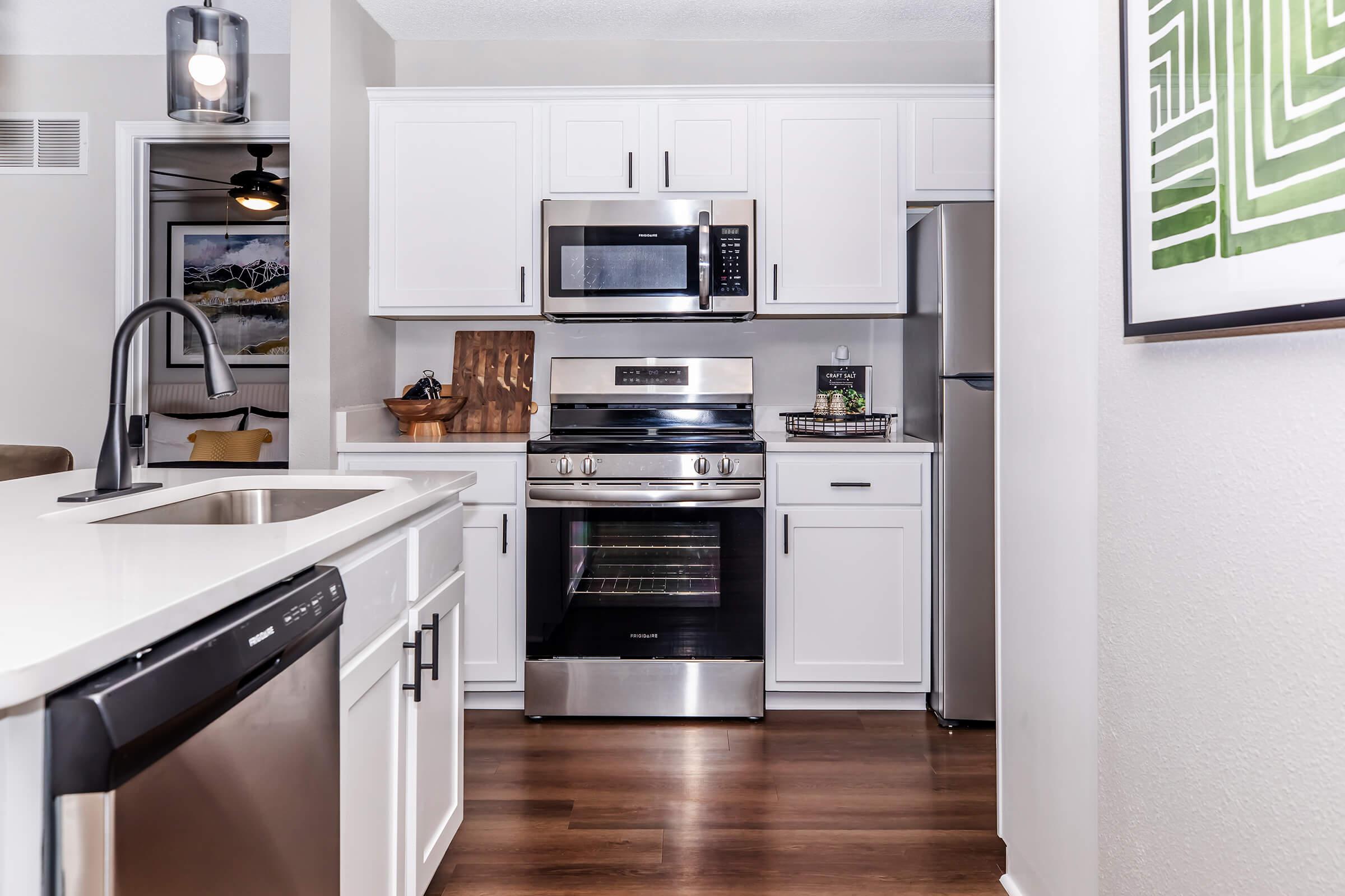 Modern kitchen featuring white cabinetry, stainless steel appliances, including a microwave and oven, and a dishwasher. The countertop is light-colored with a sink, while the flooring is hardwood. A decorative item is displayed on the counter, and there is a glimpse of a living area with artwork in the background.