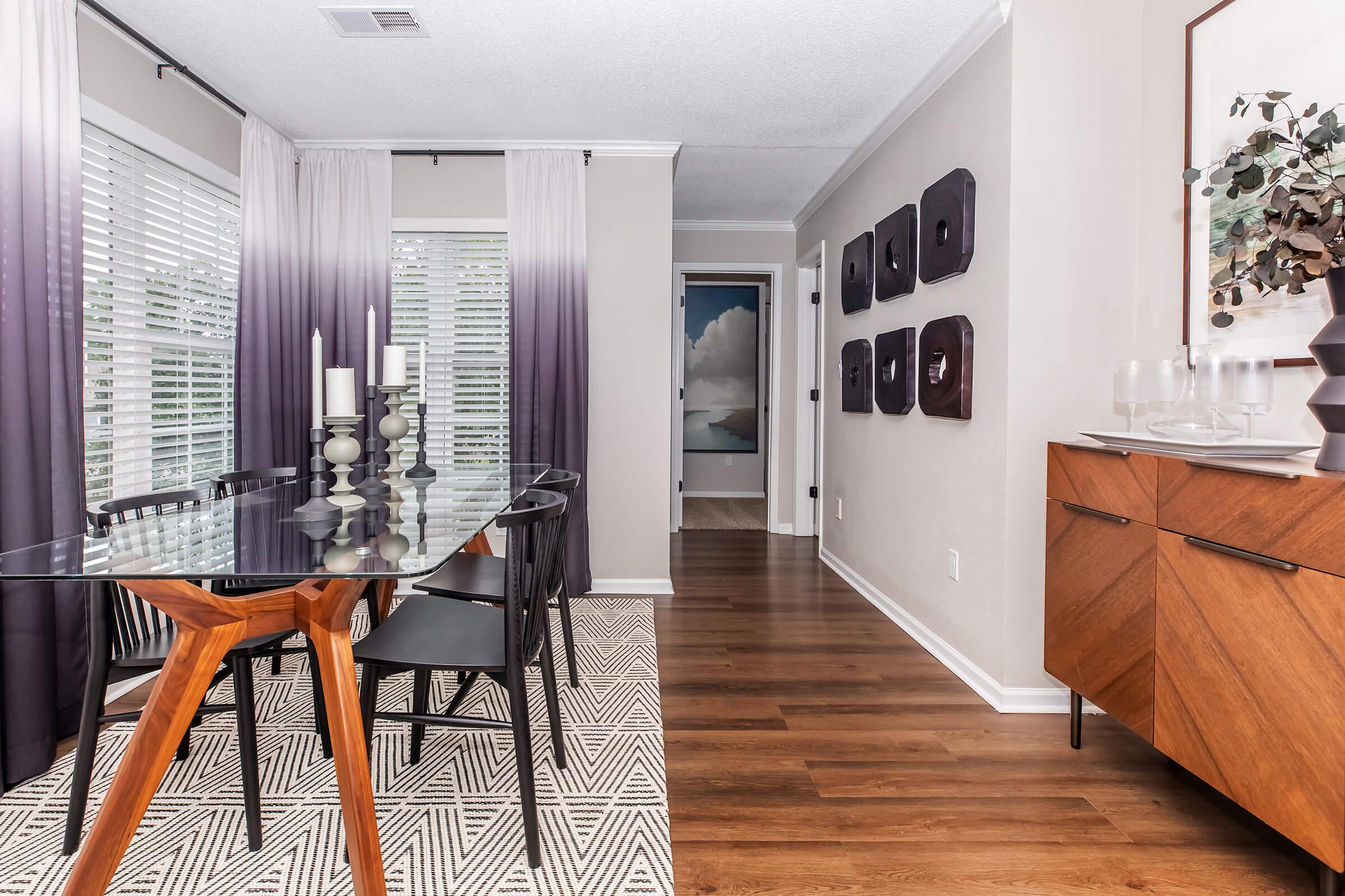 A modern dining area featuring a glass-top table with wooden legs, black chairs, and a patterned rug. Natural light filters in through sheer drapes, creating a warm atmosphere. The walls are decorated with framed art and there’s a sleek sideboard displaying glassware. A hallway leads to another room.