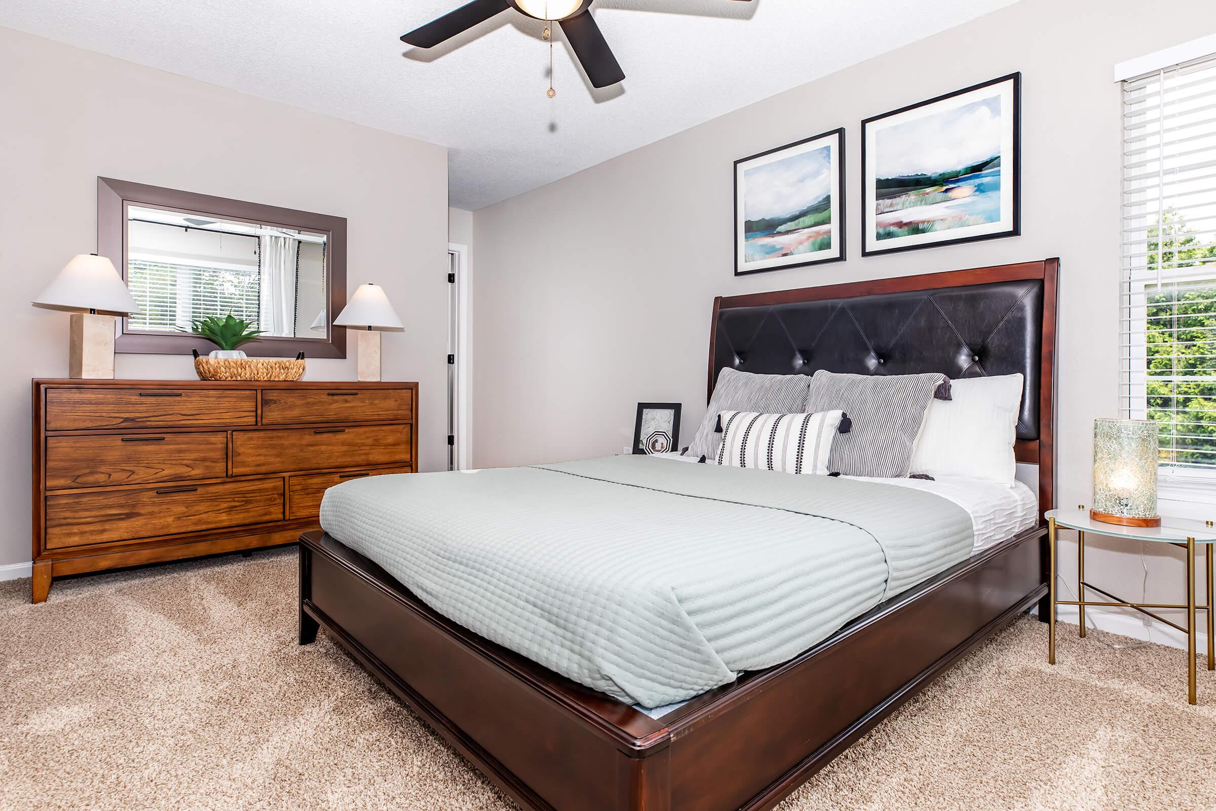 A cozy bedroom featuring a queen-sized bed with light green bedding. There's a wooden dresser with a mirror above it, two bedside lamps, and a decorative plant. The room has neutral wall colors, a ceiling fan, and framed artwork on the wall. Natural light flows through a window, adding warmth to the space.