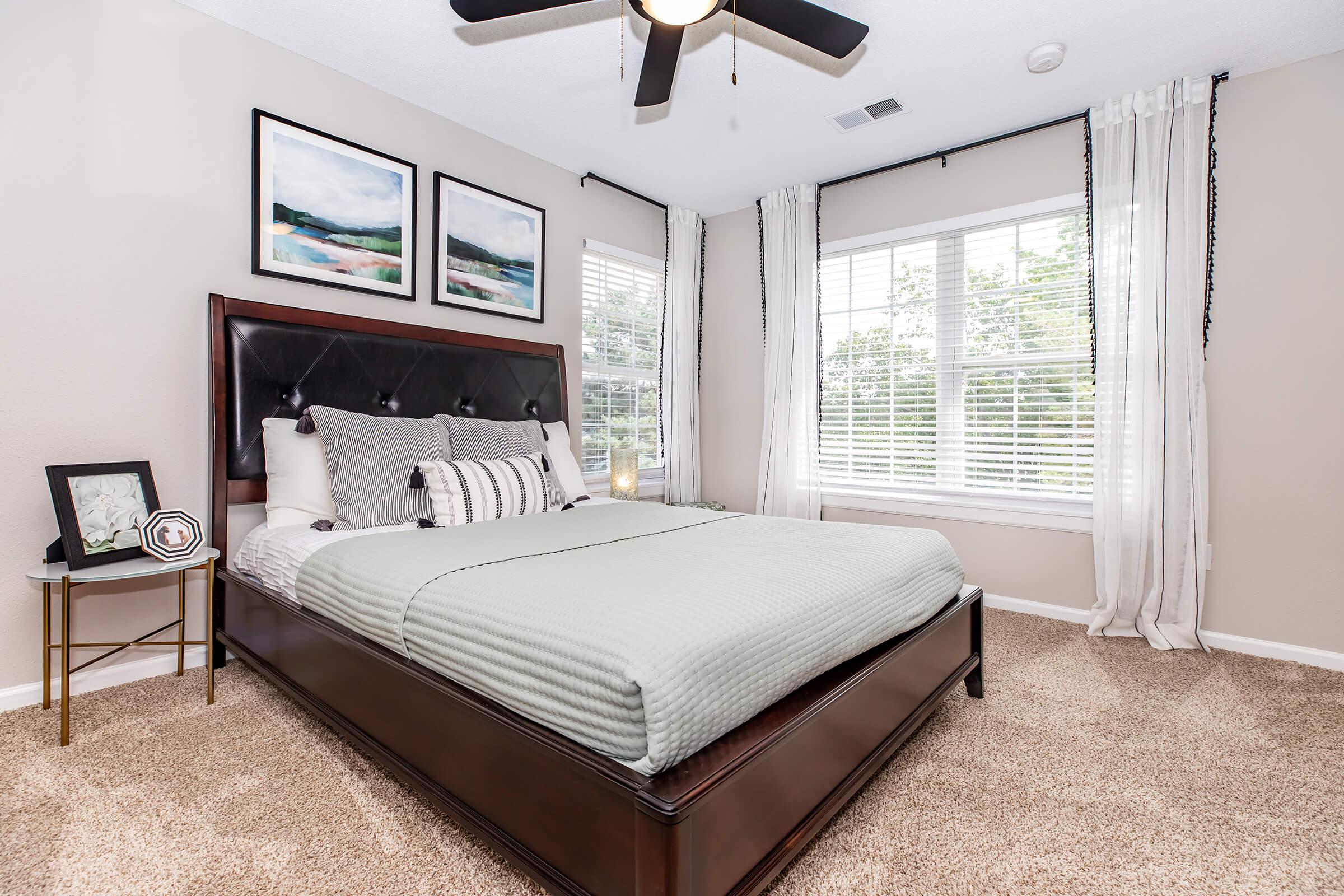 A cozy bedroom featuring a queen-sized bed with a light green cover, flanked by two decorative pillows. The room has large windows with white curtains, allowing natural light. Two framed landscape pictures hang above the bed, and there's a small side table with a decorative item. The floor is covered with light carpet.