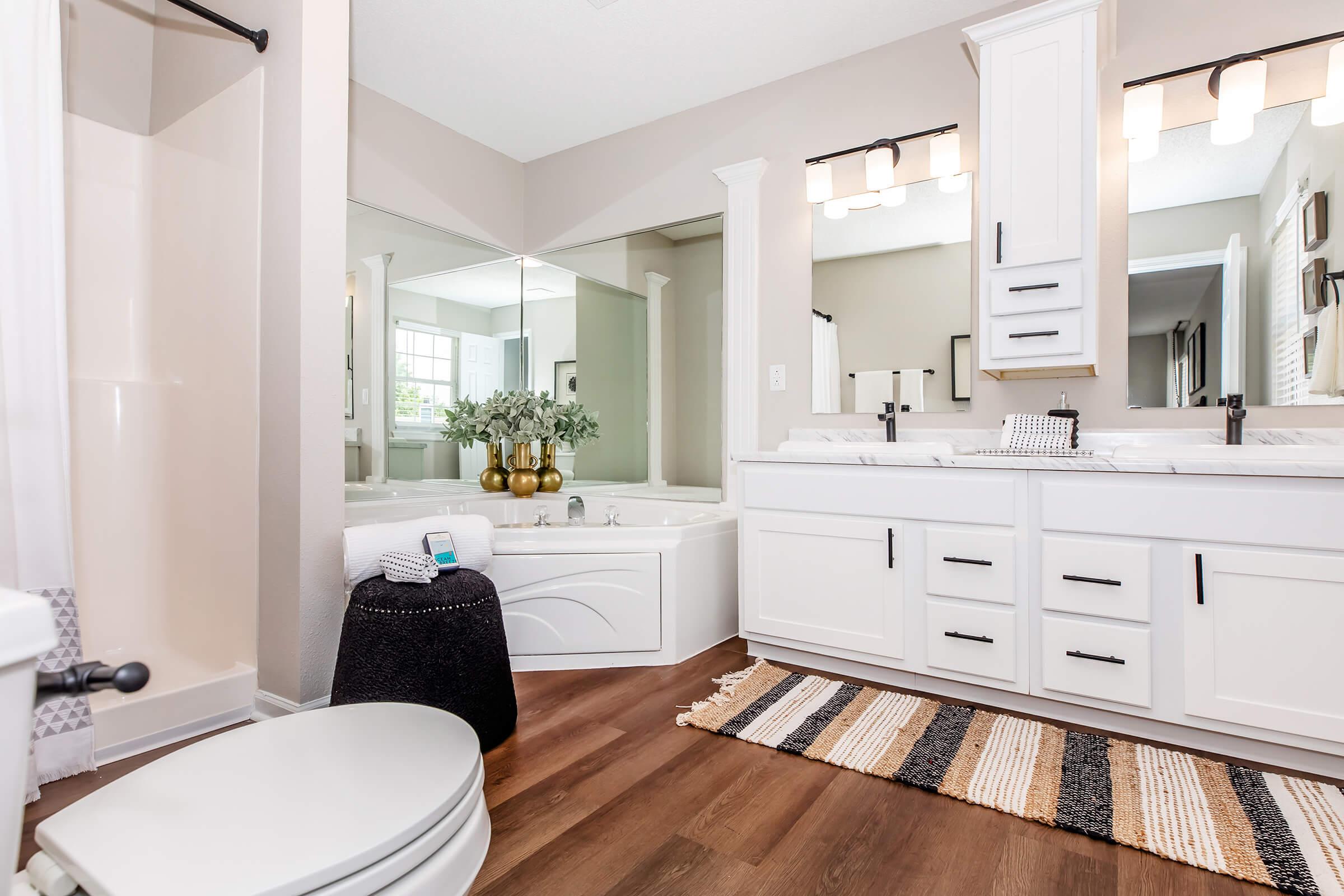 A modern bathroom featuring a double vanity with white cabinets and black hardware, a freestanding tub, and a large mirror. Stylish decor includes potted plants and a textured area rug, while natural light brightens the space through a window. A toilet and shower area are also visible.