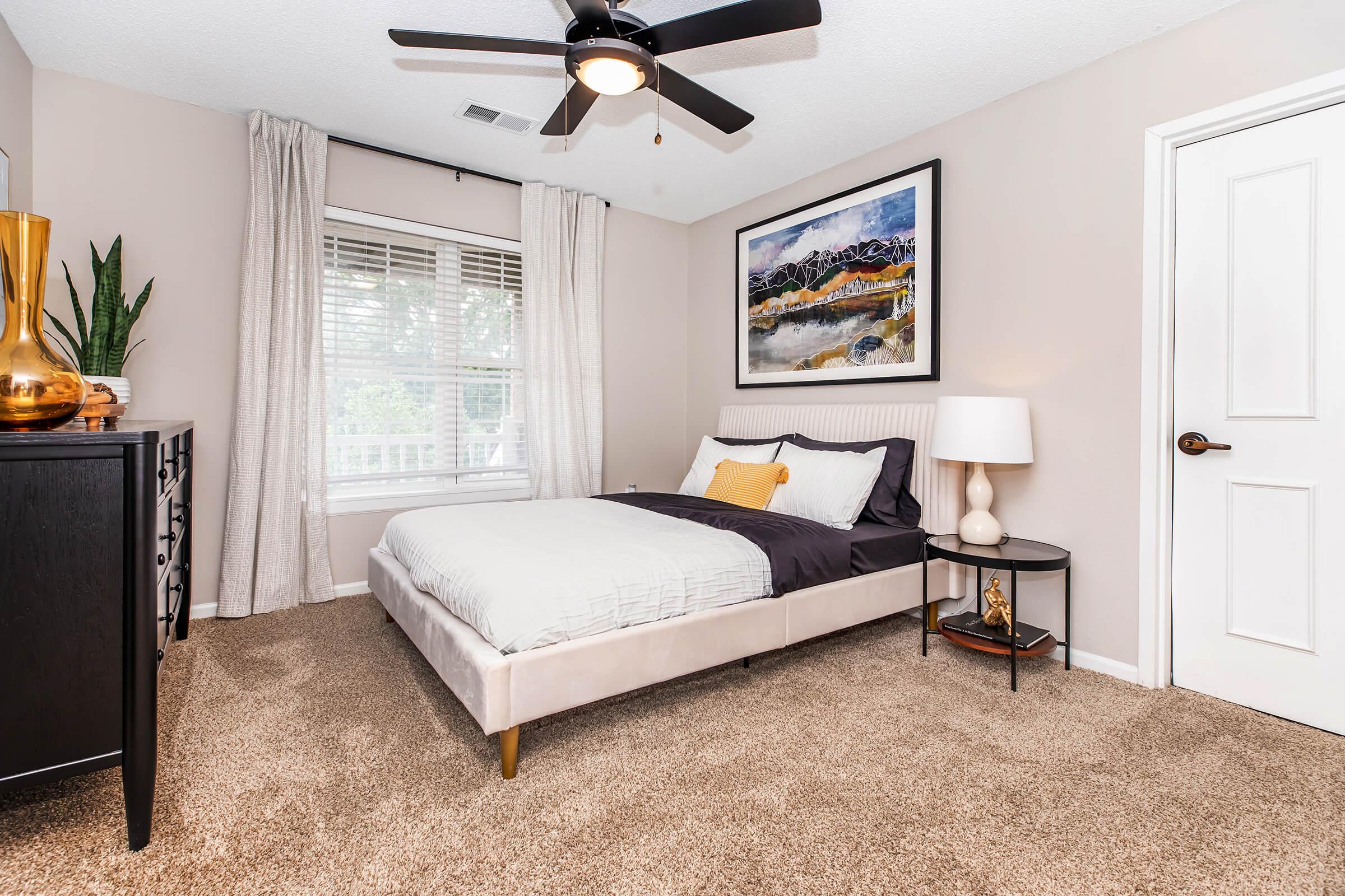 A cozy bedroom featuring a modern bed with a dark comforter and a decorative pillow. The room has a large window with sheer curtains, a stylish black dresser, and a bedside table with a lamp. A vibrant landscape painting adorns the wall, and the carpeted floor adds warmth to the space.