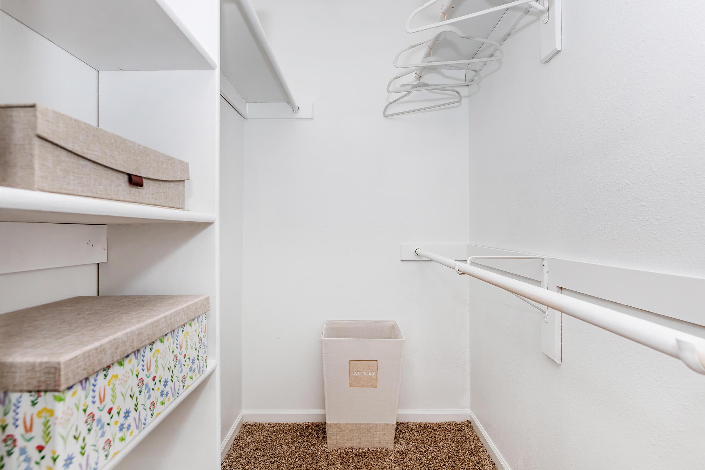 A minimalist closet interior featuring white shelves on the left, a hanging rod for clothes, a neutral-colored waste bin at the center, and two decorative storage boxes on the shelves, all set against a clean white wall and a soft carpeted floor.