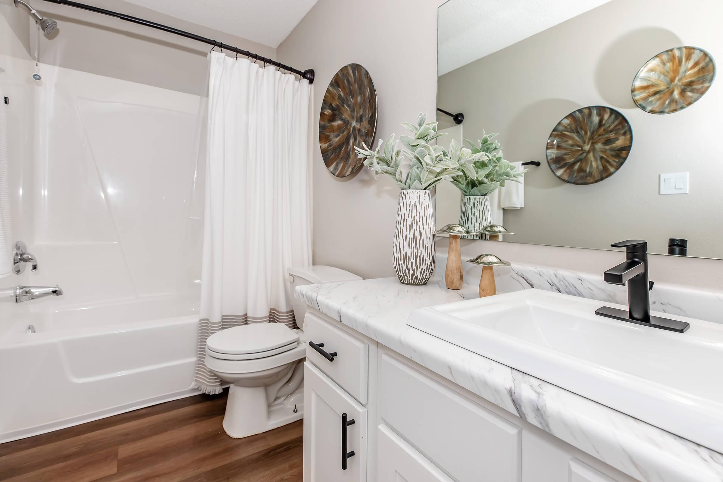 A modern bathroom featuring a white vanity with a marble countertop, a mirror, and decorative elements. There is a bathtub with a shower curtain, a toilet, and decor such as potted plants and wall mirrors. The flooring is wooden, adding warmth to the space.