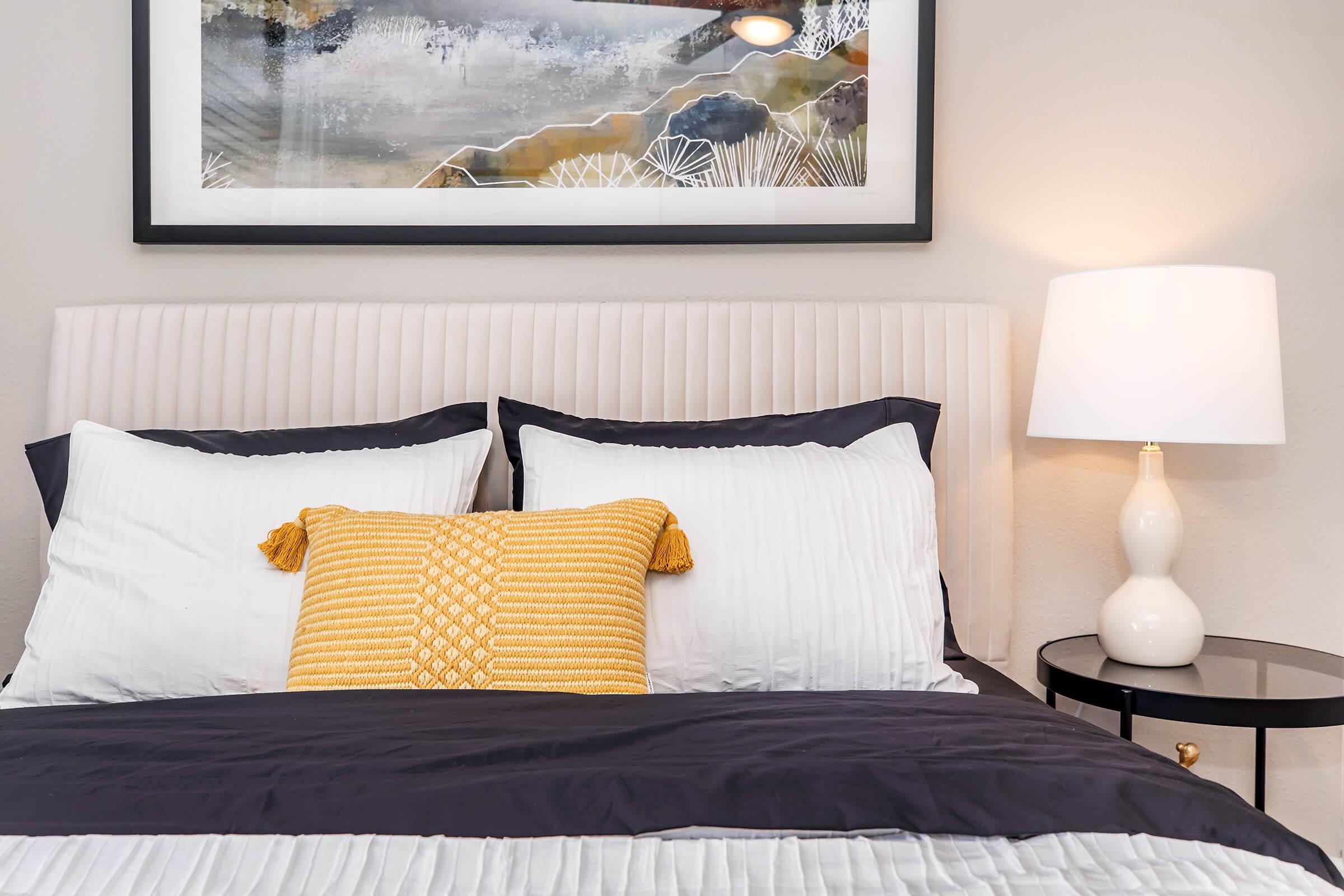 A well-decorated bedroom featuring a light-colored headboard with two white pillows and a yellow decorative pillow. The bedspread is dark blue, and beside the bed is a round black side table with a white lamp. A framed artwork hangs above the bed, showcasing abstract landscape elements.