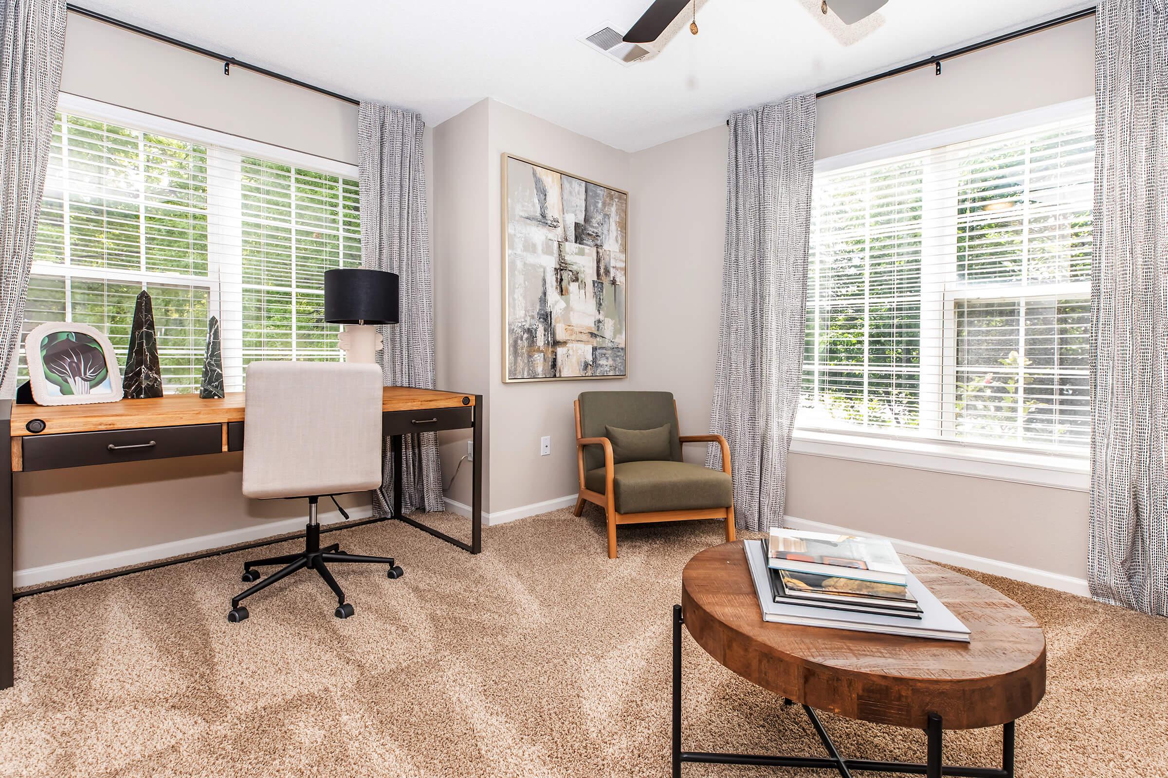 A modern home office featuring a wooden desk with a chair, a brown accent chair, and a round coffee table. The room is adorned with large windows covered by sheer curtains, allowing natural light to illuminate a colorful abstract painting on the wall. The carpet is light and neutral-toned.