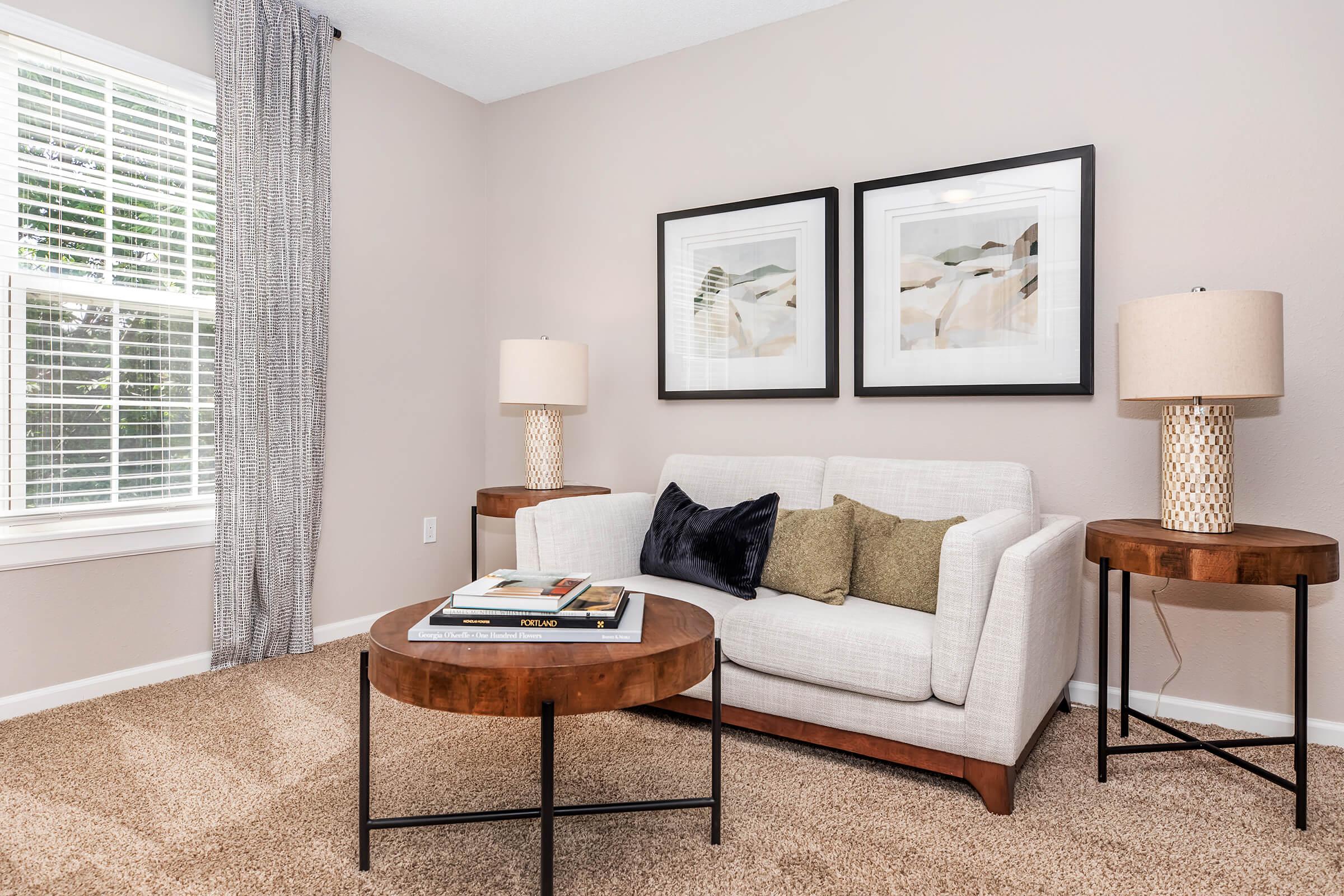 A cozy living room featuring a light-colored sofa with pillows, two contemporary side tables, and elegant lamps. Above the sofa are two framed artworks. There's a large window with sheer curtains letting in natural light, and the room has a warm, inviting carpet.