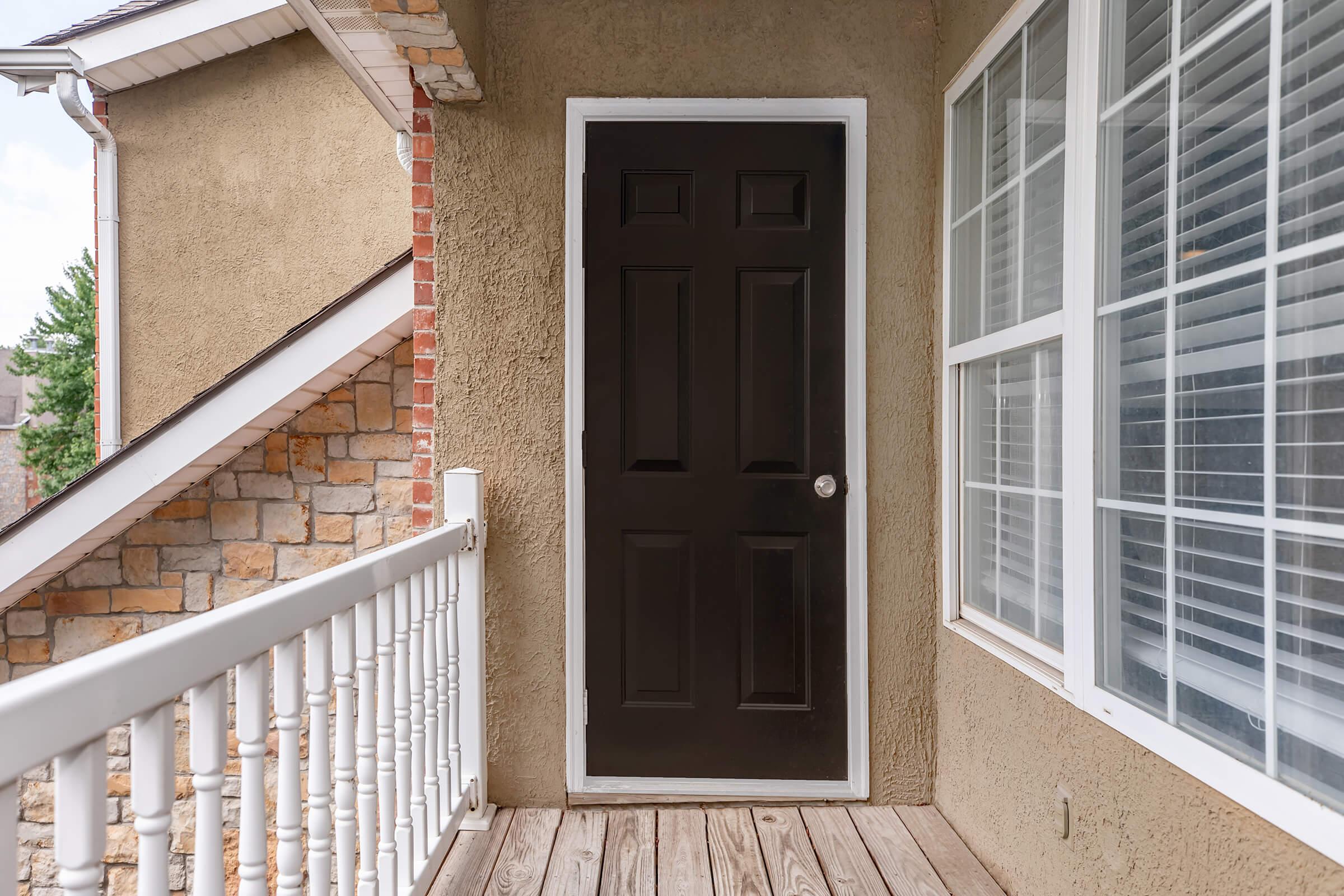 A black front door surrounded by a textured beige wall, with a white railing and wooden deck. To the side, there's a window with multiple panes, letting in natural light, and a staircase leading to a higher level in the background.