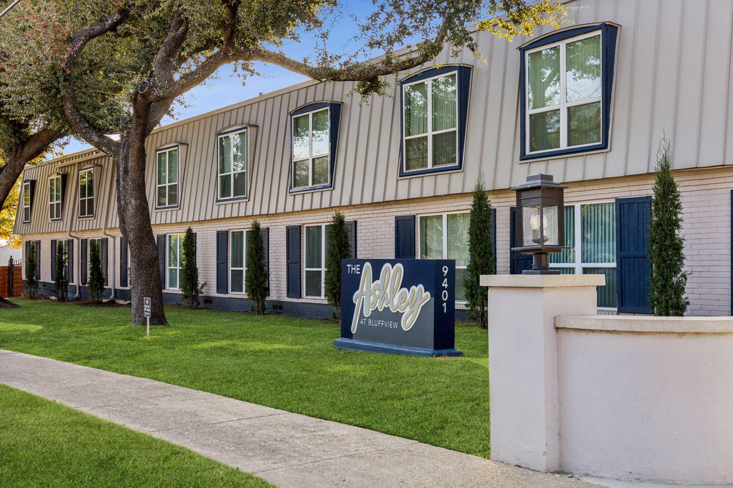 Multi-story apartment building with a modern exterior, featuring large windows and a sign reading "The Ashley" at the entrance. The building is surrounded by well-maintained grass and trees, creating a welcoming atmosphere. The address "9401" is visible on the sign.