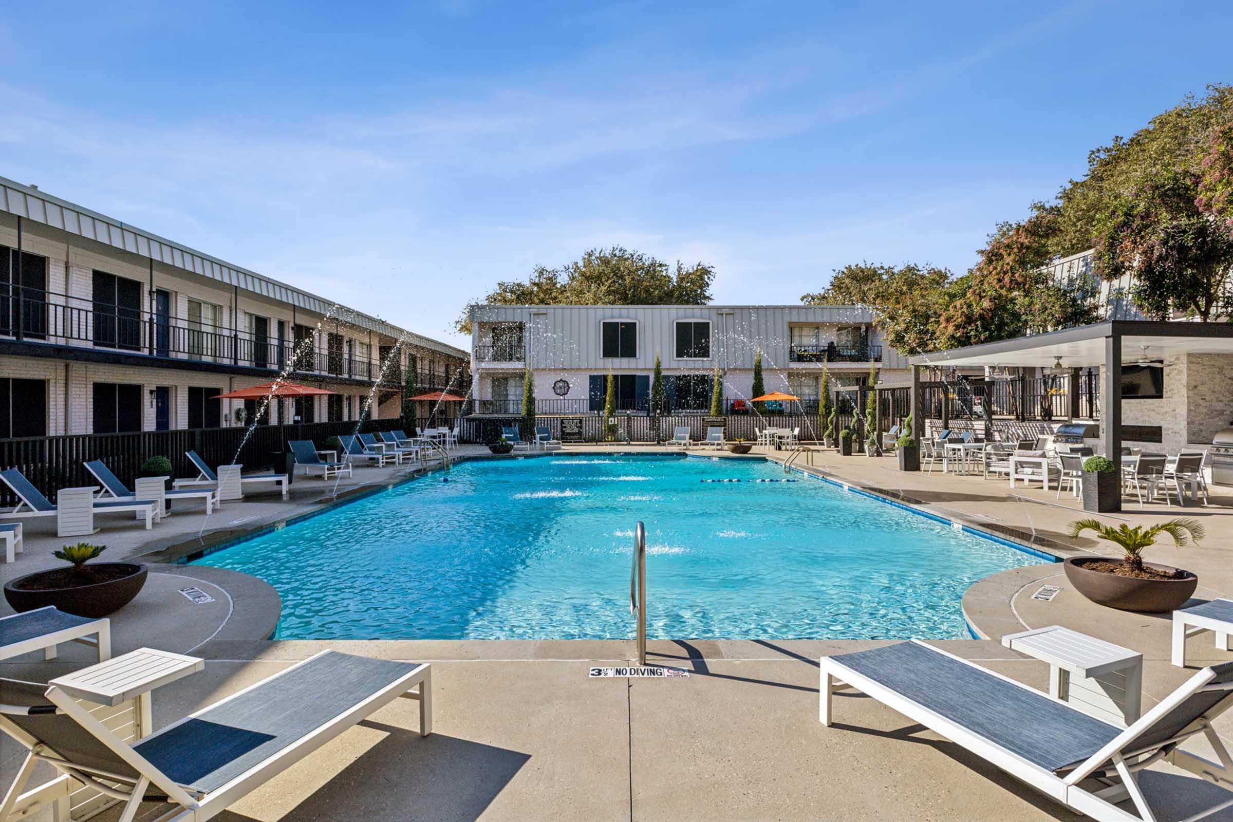 A clear blue swimming pool surrounded by lounge chairs and shaded seating areas. Nearby buildings feature balconies and outdoor decorations, with trees providing a touch of greenery in the background under a bright blue sky.