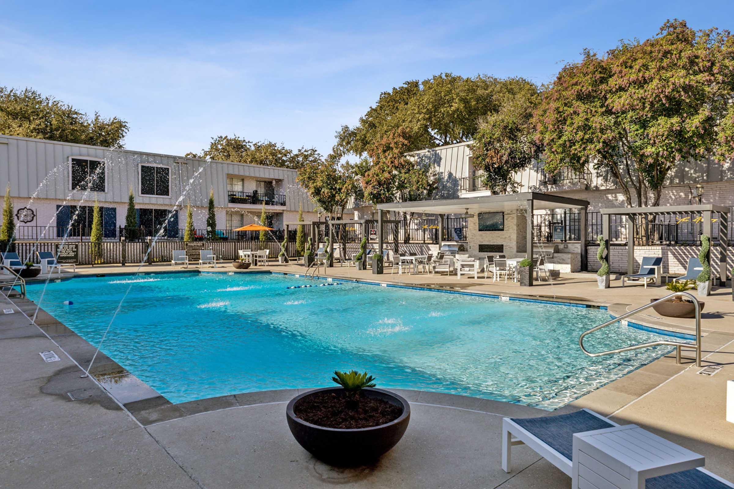 A spacious outdoor swimming pool surrounded by lounge chairs and tables. Fountains spray water into the pool. Lush greenery and trees are visible in the background, along with modern residential buildings. Sun loungers and shaded areas create a relaxing atmosphere for leisure and socializing.