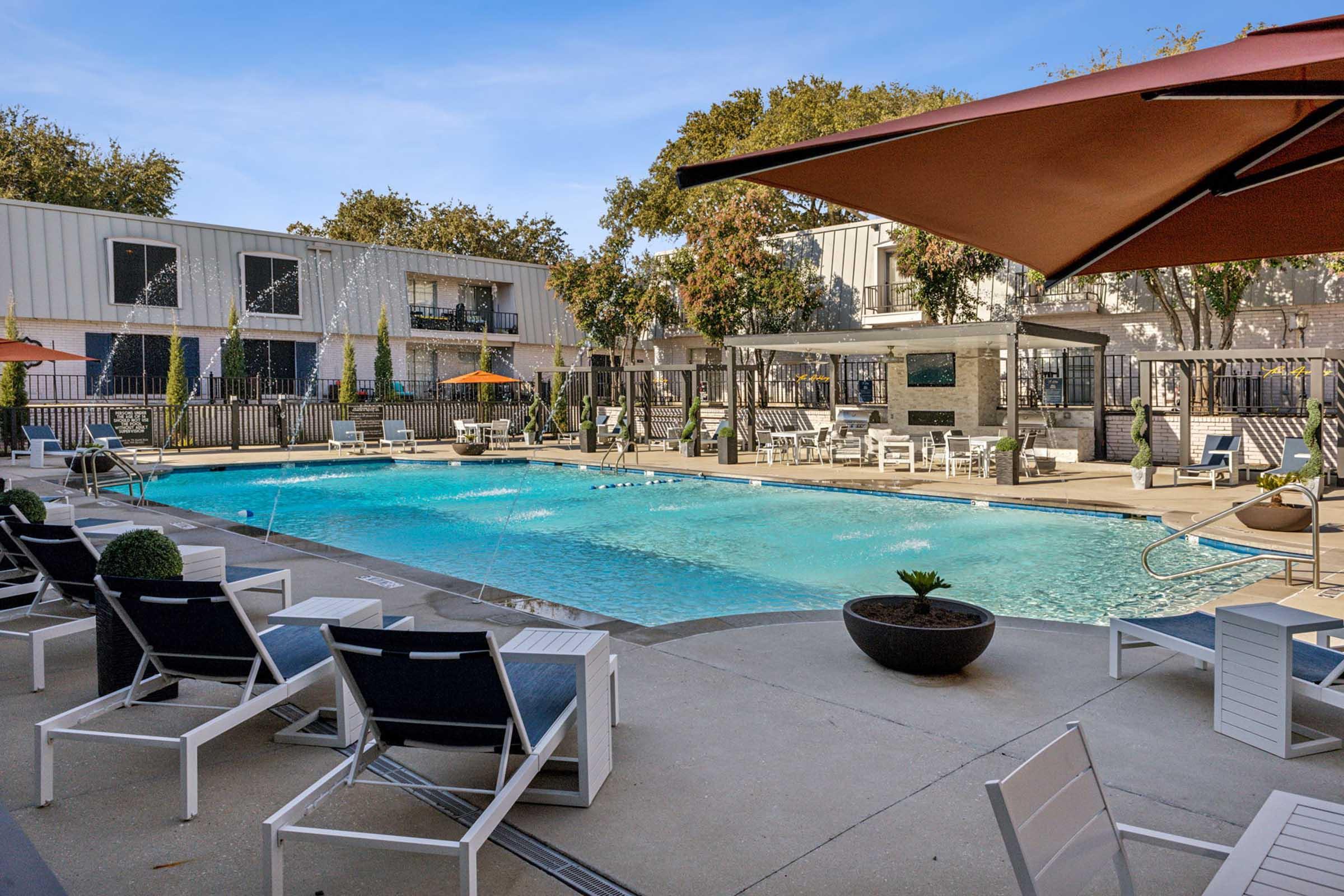 A bright outdoor swimming pool surrounded by sunny lounge chairs and umbrellas. Lush greenery and modern buildings are visible in the background, creating a relaxing atmosphere. The pool area features a contemporary design with various seating arrangements for guests to enjoy.