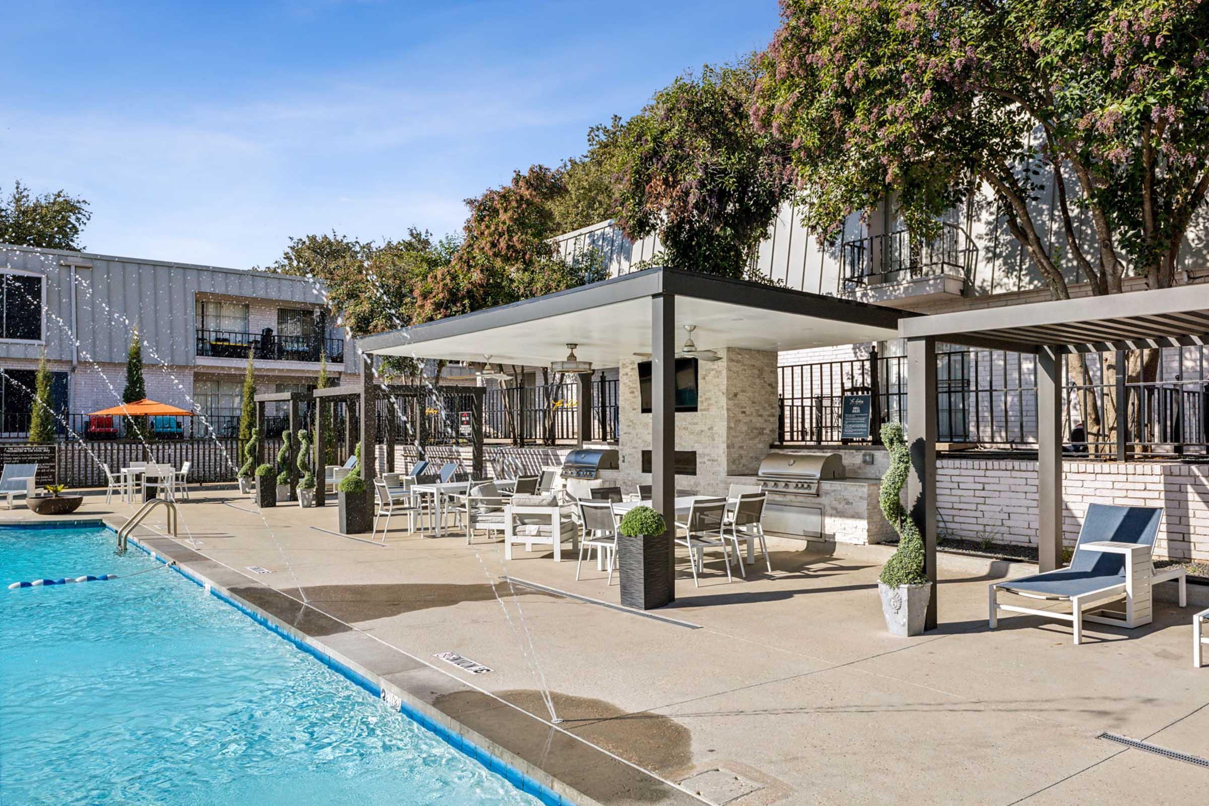 A bright and inviting pool area featuring a clear blue pool surrounded by lounge chairs and shaded seating. There are potted plants and a covered grilling station under a pergola. In the background, there are two-story apartment buildings set amidst trees, creating a relaxed outdoor atmosphere.
