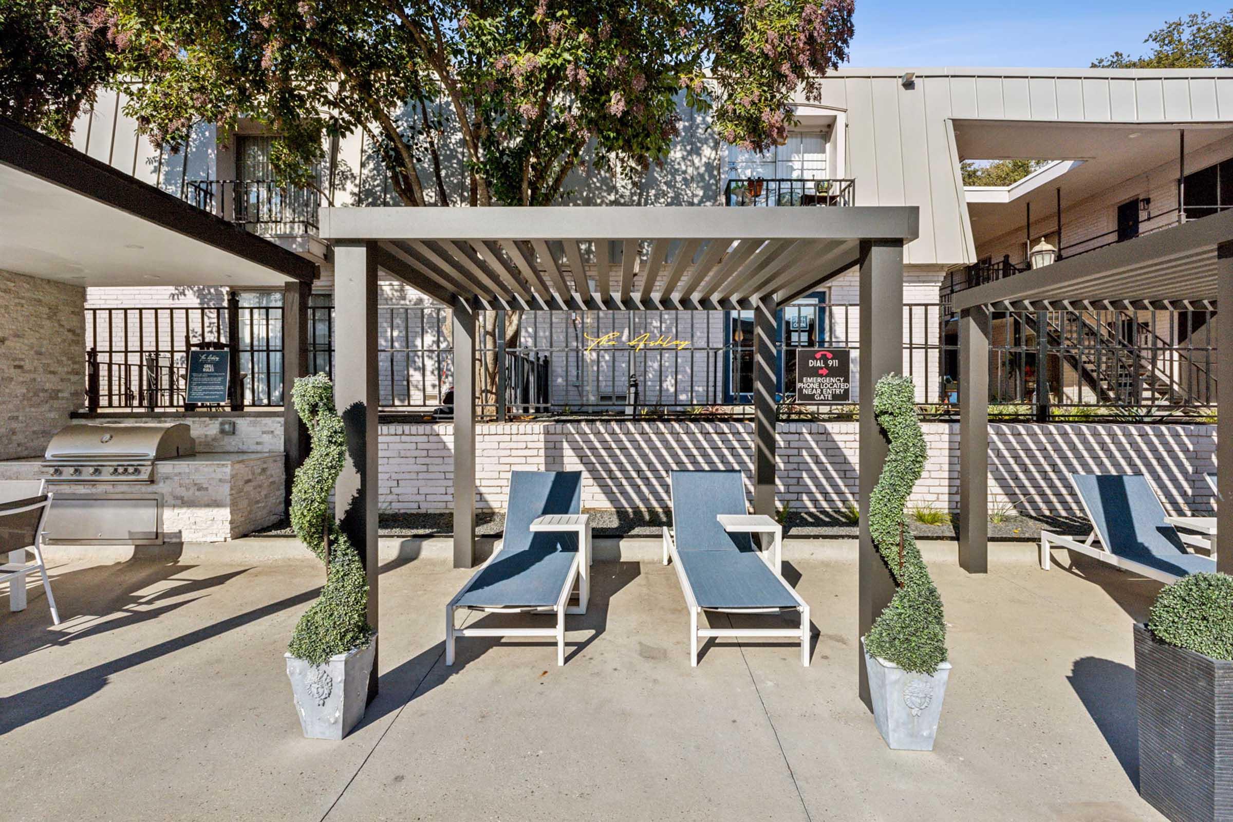 A poolside area featuring two lounge chairs under a pergola, flanked by decorative green topiary. In the background, a barbecue grill and a multi-story building with an outdoor seating area are visible, set against a clear blue sky and lush trees.