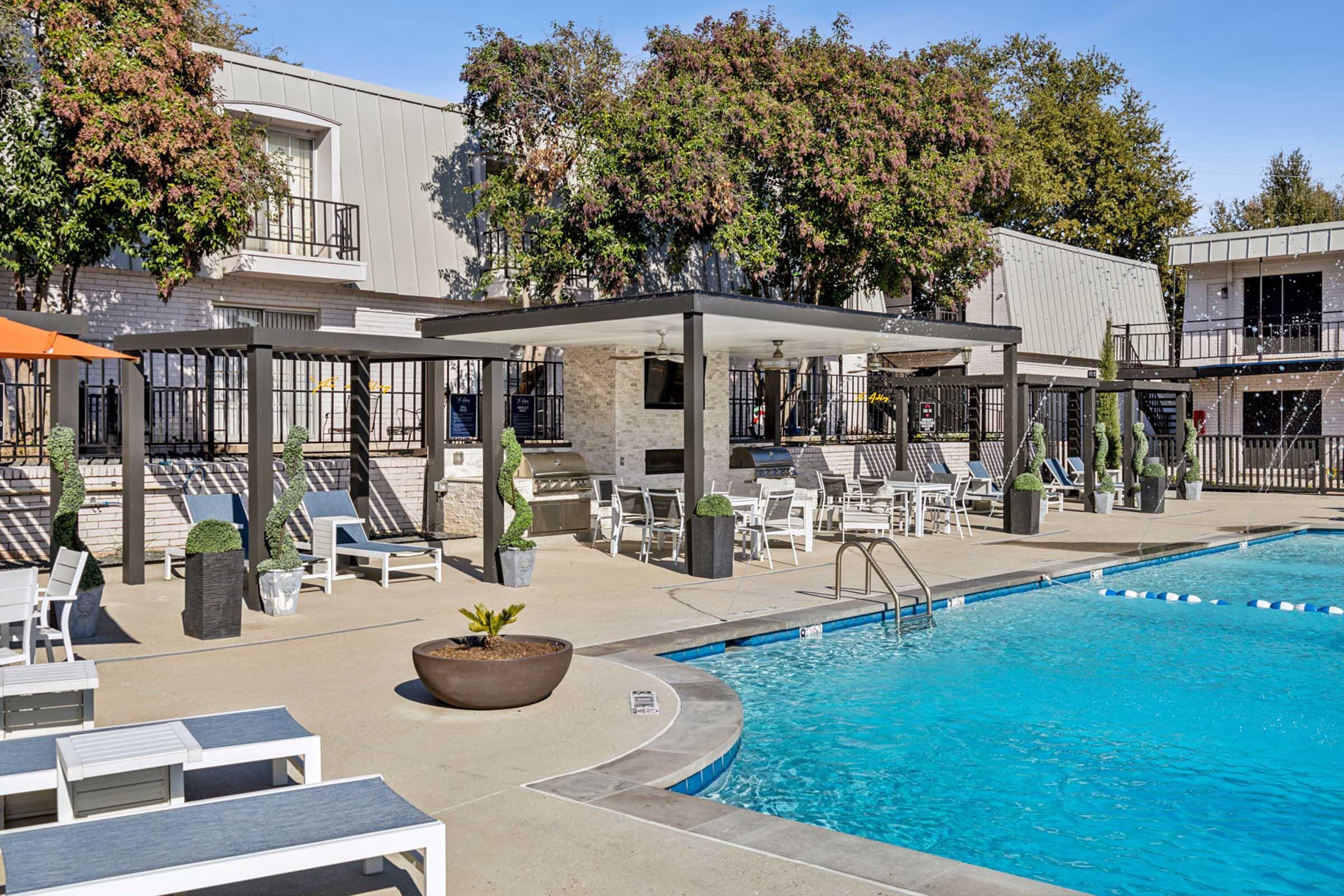 A sunny pool area featuring a clear blue pool, lounge chairs, and cabanas. Surrounding the pool are well-maintained trees and potted plants, along with barbecue grills and outdoor seating. The area is bordered by fencing and nearby buildings, creating a relaxing atmosphere for relaxation and socializing.