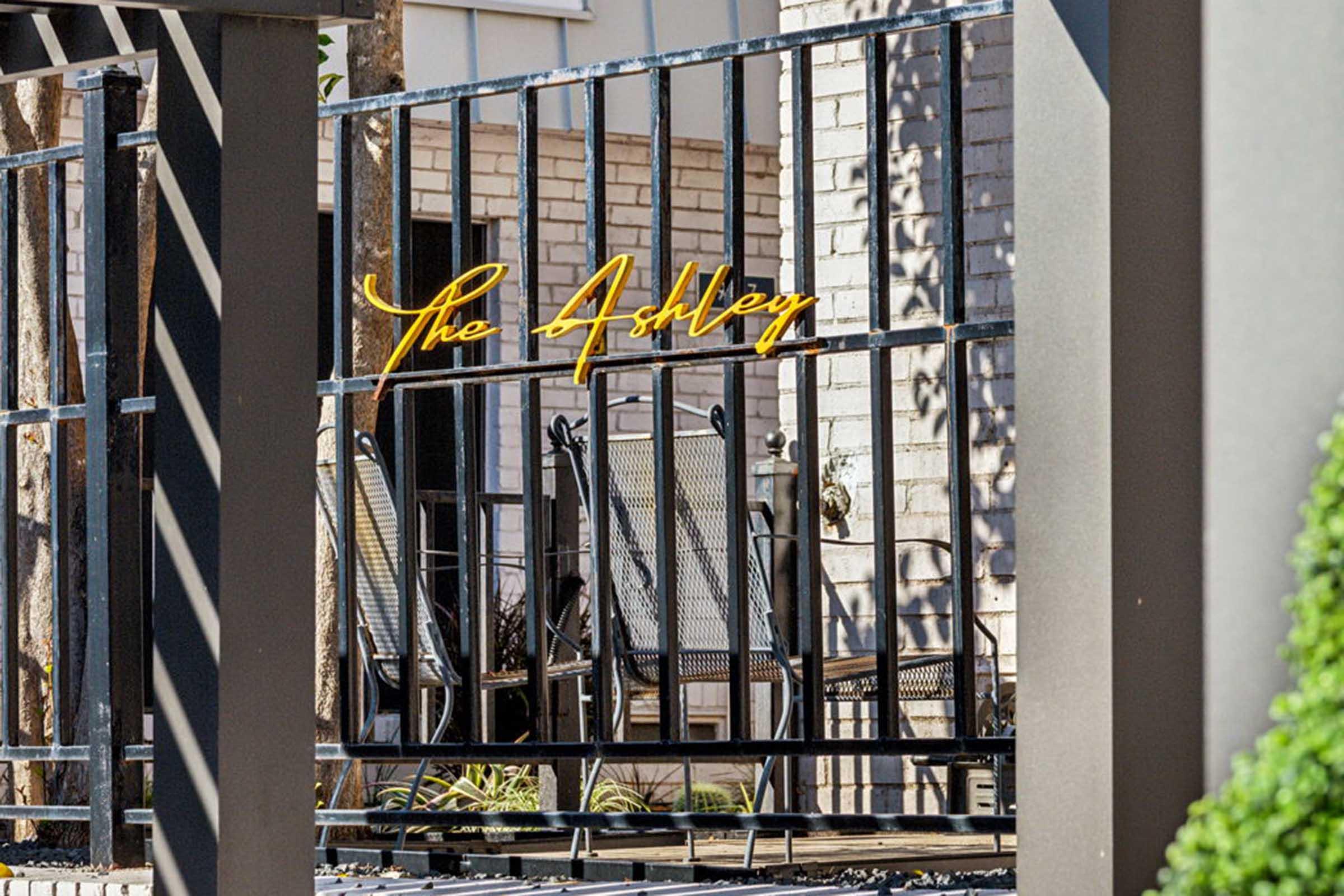 A decorative gate with the text "The Ashley" in yellow lettering, framed by a modern black metal fence. Behind the gate, there are two comfortable chairs and a small area with greenery, suggesting a welcoming atmosphere for a residence or establishment.