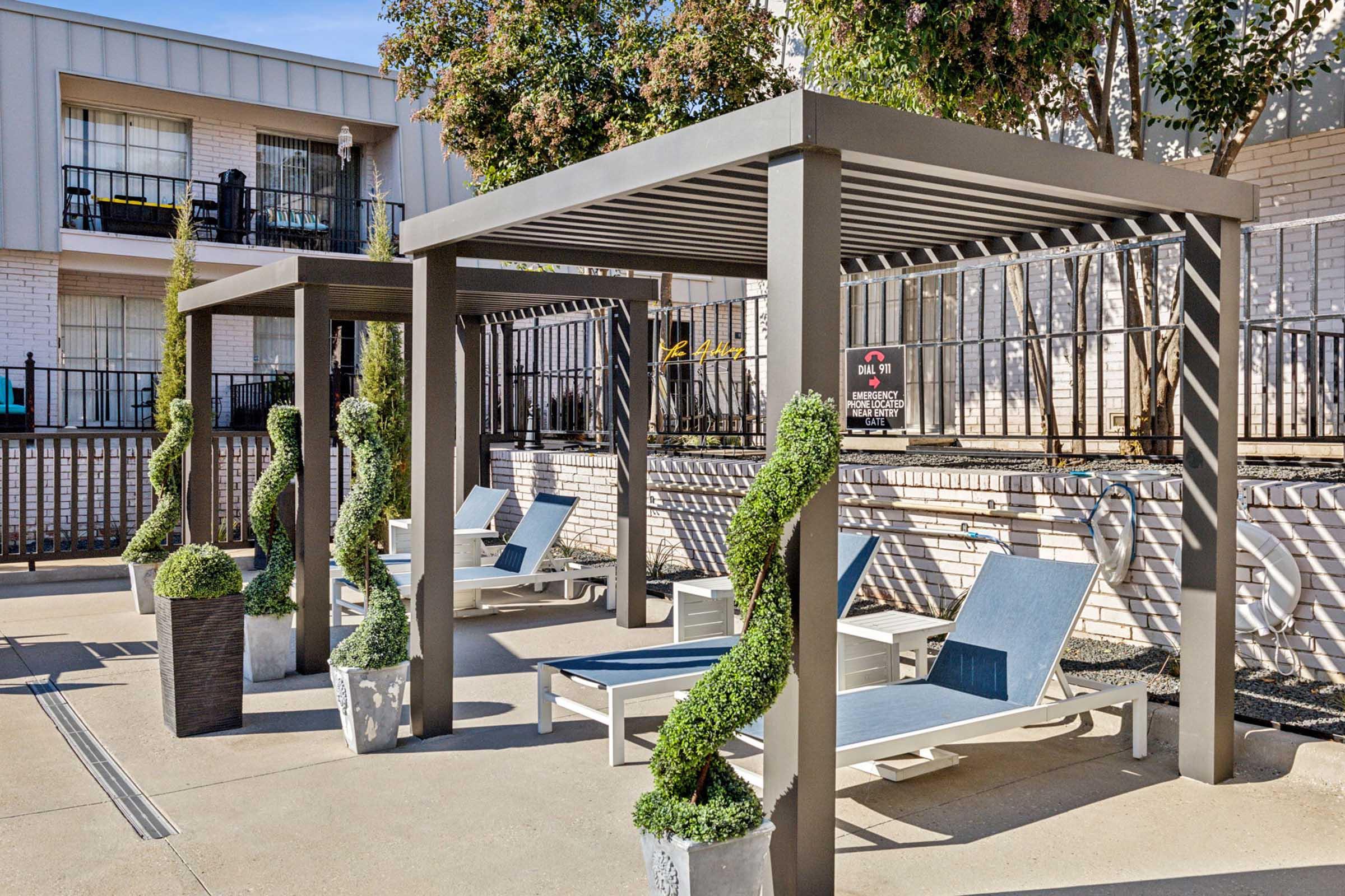 Two shaded pergolas with lounge chairs underneath, surrounded by neatly trimmed greenery in planters. The area is well-maintained, featuring a poolside environment with a fence in the background. Sunlight casts shadows on the concrete surface, creating a relaxing outdoor space.