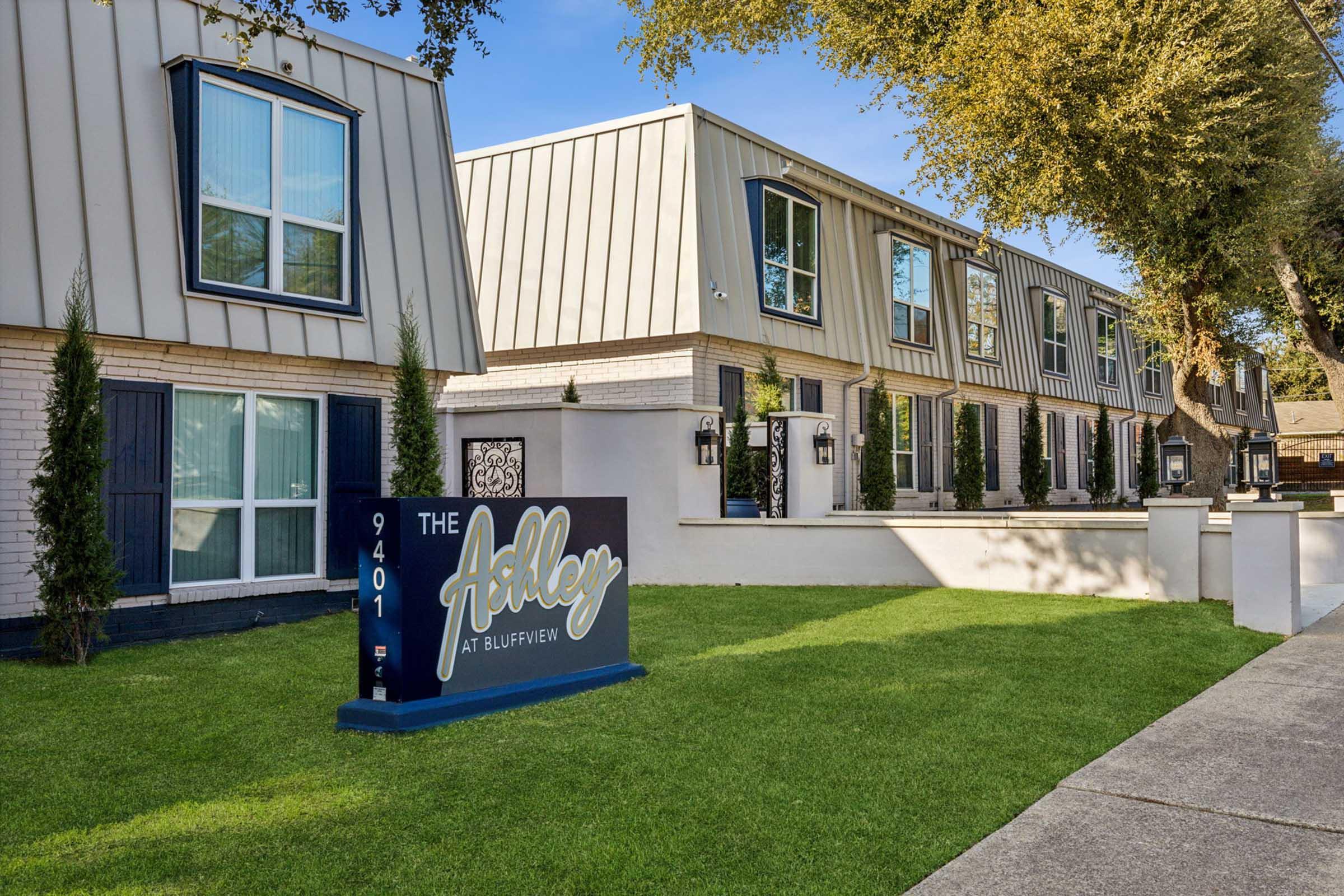 A well-maintained apartment complex featuring modern architecture. The building has multiple units with large windows and is surrounded by a neat lawn. A prominent sign displays "The Ashley at Bluffview" along with an address. The sky is clear, and there are trees nearby, enhancing the landscaped surroundings.