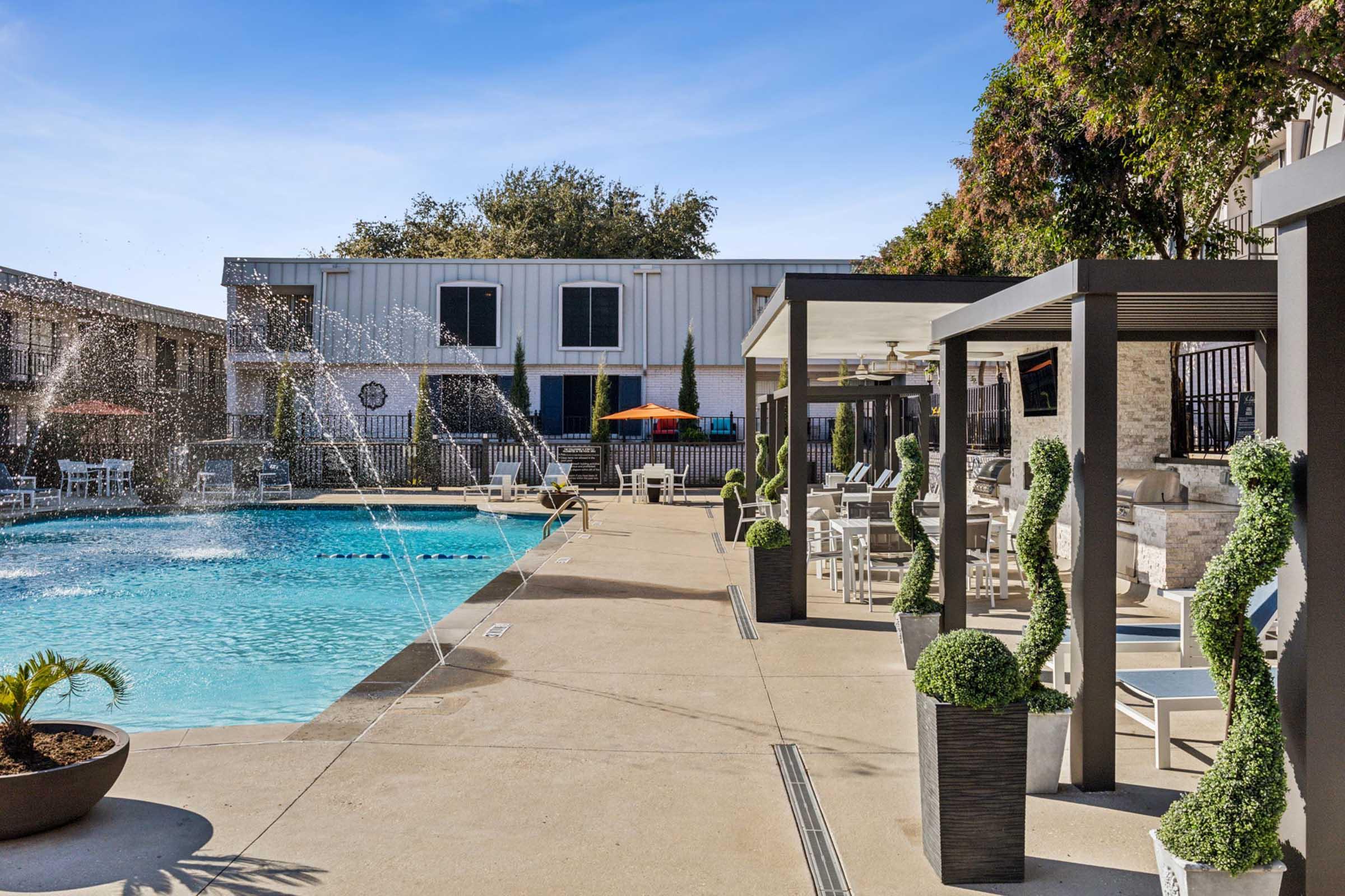 A bright and inviting pool area featuring a clear blue pool with water fountains, surrounded by lounge chairs and potted plants. Cozy seating areas with modern decor are visible, and an umbrella provides shade. Trees and buildings are in the background, creating a relaxing atmosphere.