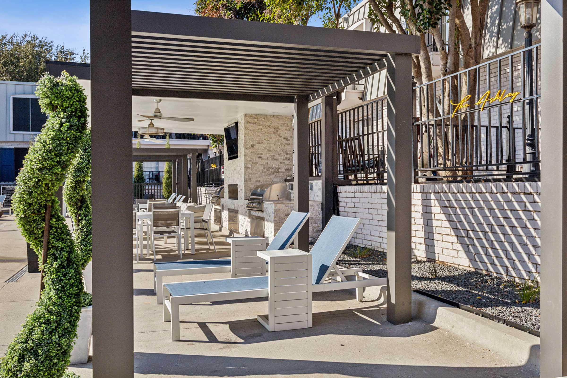 A shaded outdoor area featuring lounge chairs and a communal grill. The space is surrounded by greenery and has a modern design, with a stone wall and a flat-screen TV mounted nearby. Sunlight casts shadows on the ground, creating a relaxing ambiance for gatherings or leisure time.
