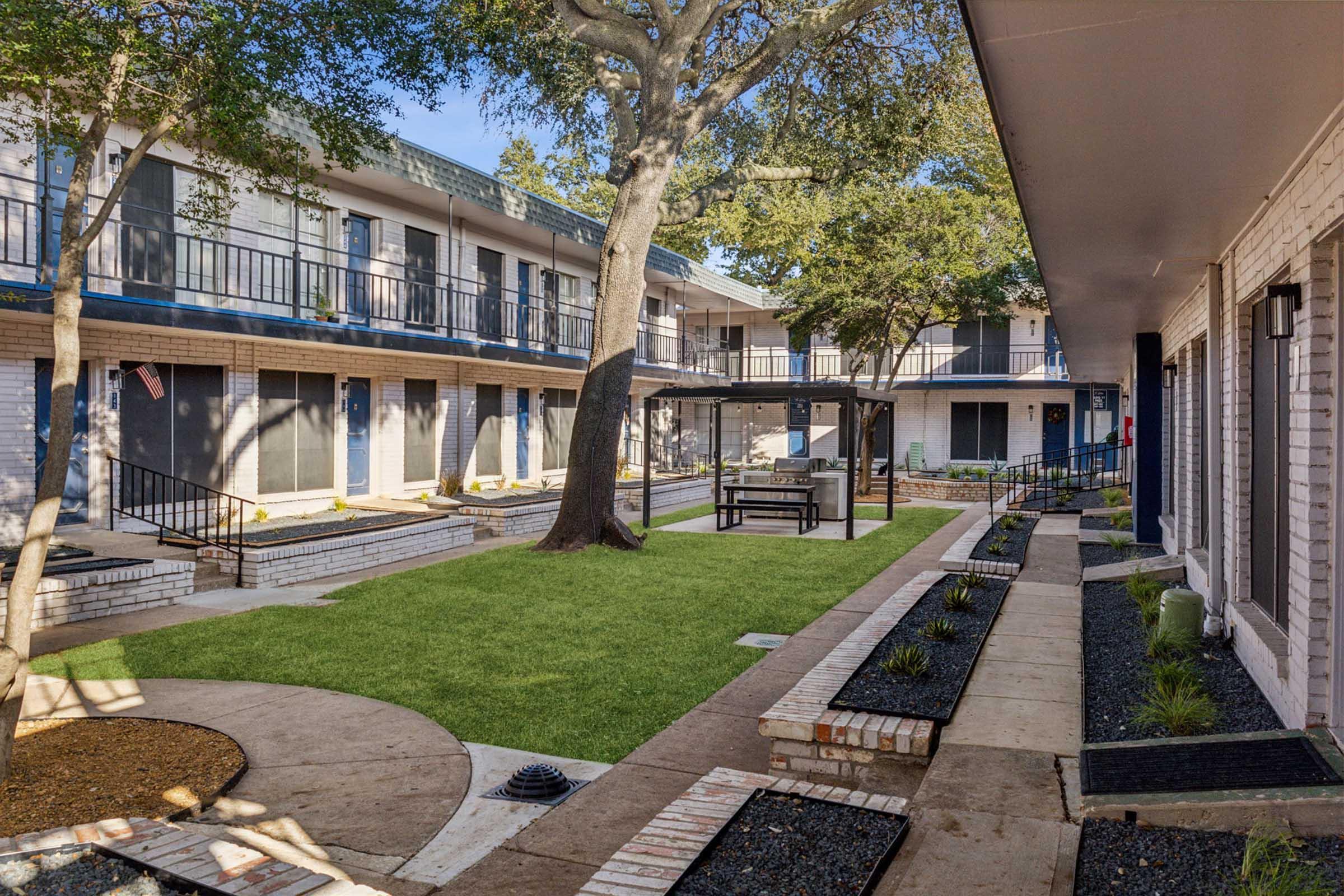 A view of a courtyard in a hotel or apartment complex featuring greenery, seating under a gazebo, and surrounding walkways. There are two-story buildings with balconies on either side, and well-maintained landscaping including grass and shrubs.