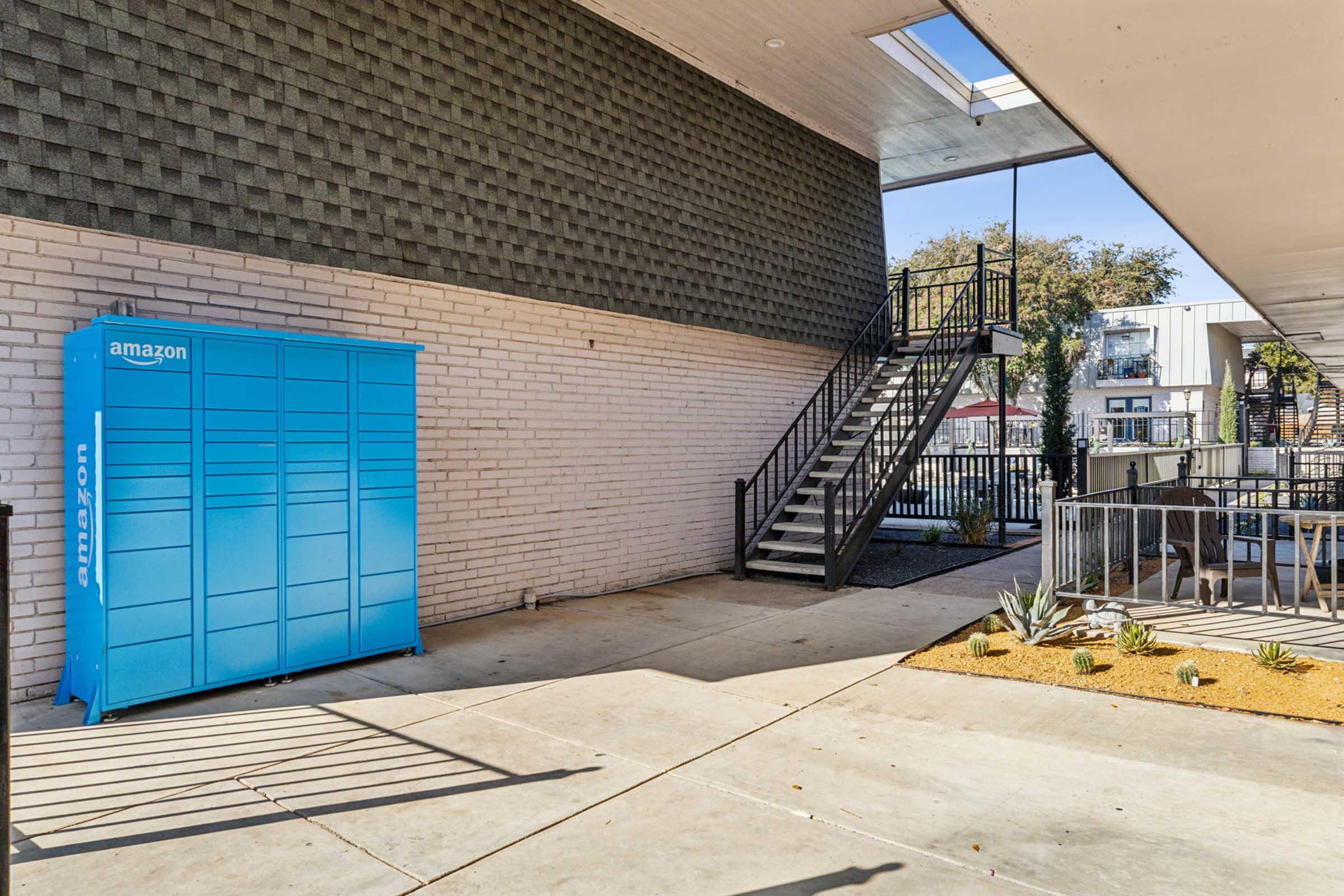 A blue Amazon locker located in an outdoor area next to a staircase. The surrounding space features concrete flooring, a small garden with plants, and a nearby building. The area is well-lit and designed for accessibility.