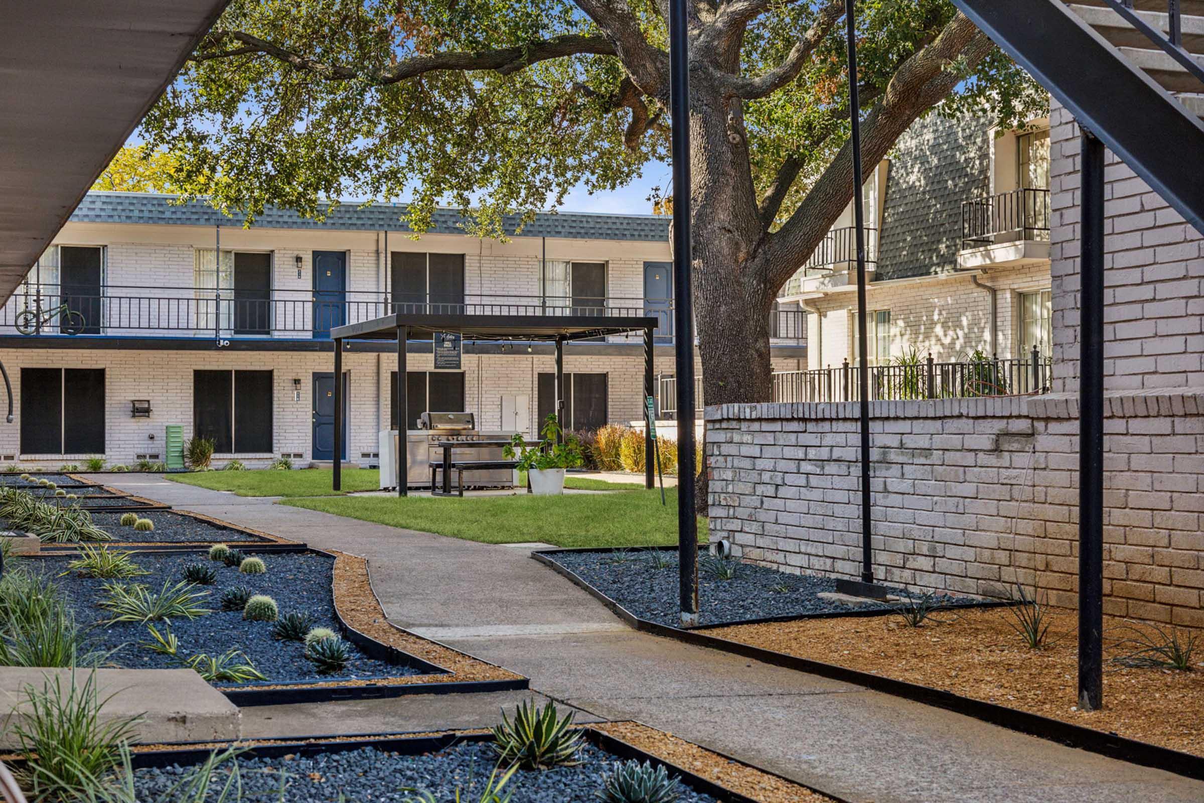 A well-maintained courtyard of an apartment complex featuring a grassy area, outdoor grilling station, and landscaped paths with small plants and rocks. The buildings are two stories with balconies, surrounded by mature trees, providing a tranquil outdoor space for residents.