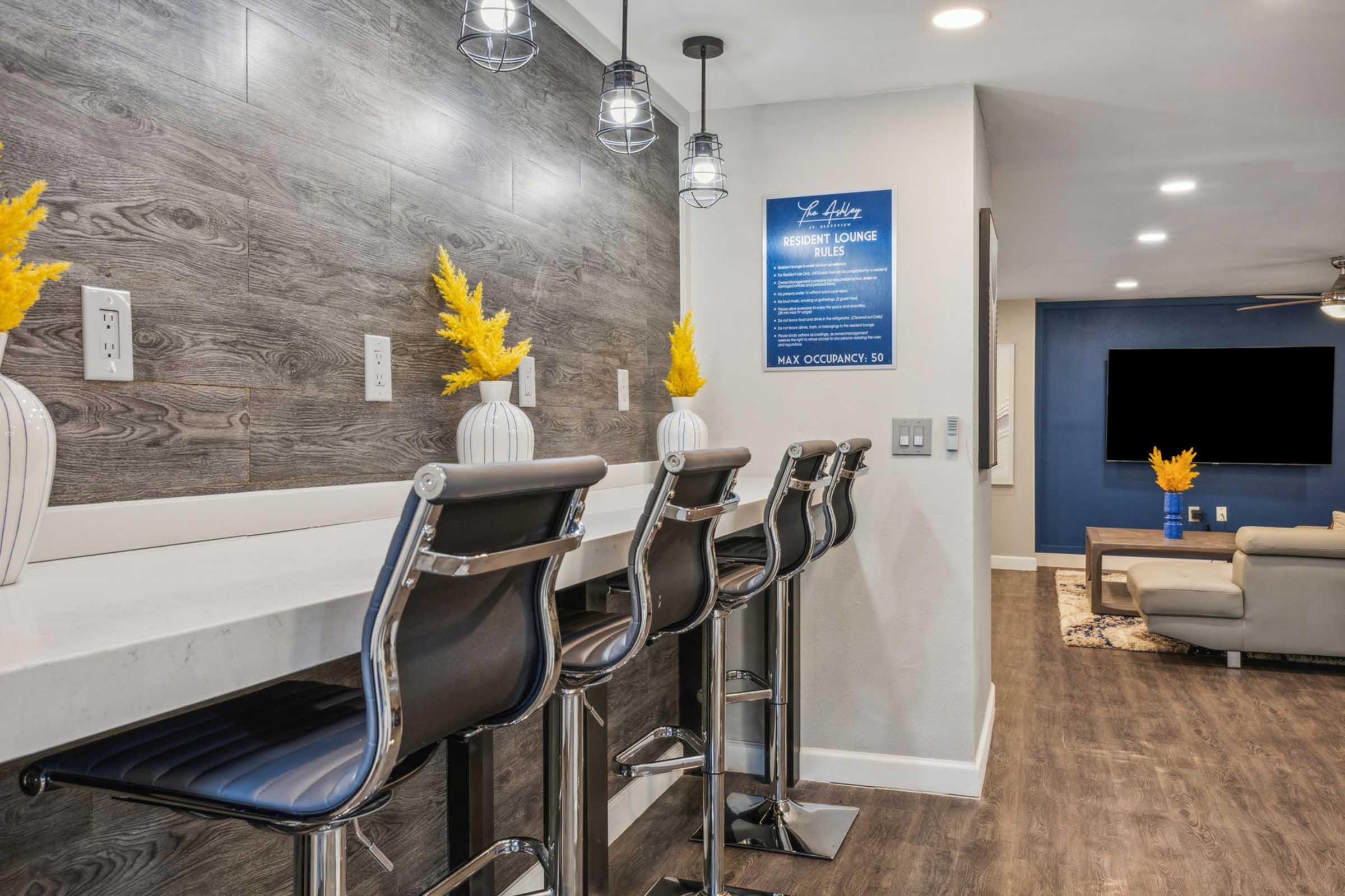 A modern lounge area featuring a bar with high-backed stools and a sleek countertop. There are decorative vases with yellow flowers on the bar, and a blue wall with information displayed. A cozy seating area is visible in the background, facing a large flat-screen television. The space has a contemporary design.