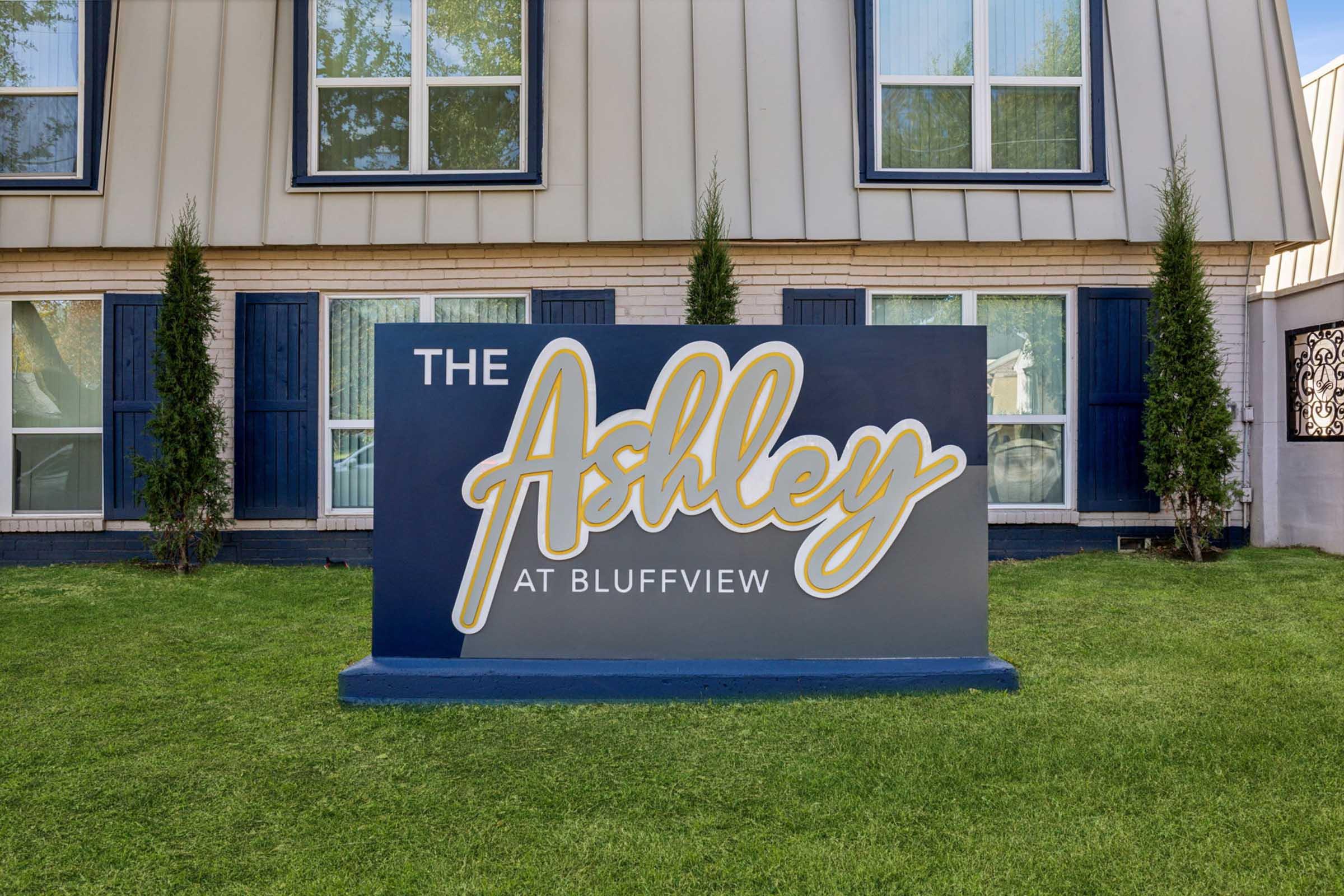 Sign displaying "The Ashley at Bluffview" in a stylish cursive font, set against a modern building facade with large windows and green landscaping.