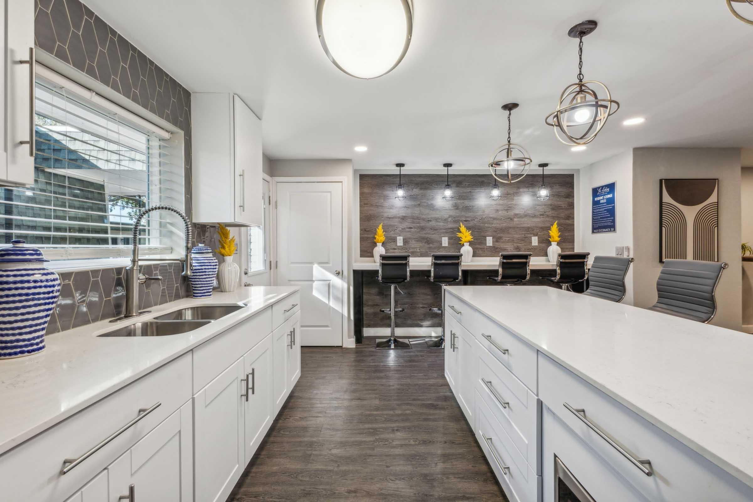 A modern kitchen featuring white cabinetry, a large island with a sleek countertop, and pendant lighting. The backsplash includes decorative tiles, while a window allows natural light. Bar stools are arranged at a counter with stylish decor, highlighting a contemporary design aesthetic.