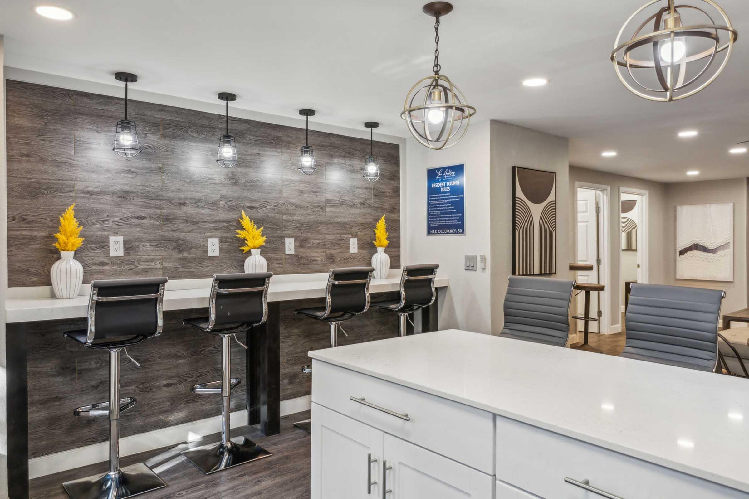 Modern kitchen with a sleek design featuring a white countertop and high bar stools. The background showcases a stylish wall with pendant lights and decorative vases with yellow flowers, enhancing the contemporary aesthetic. Suitable for a cozy dining or social area.