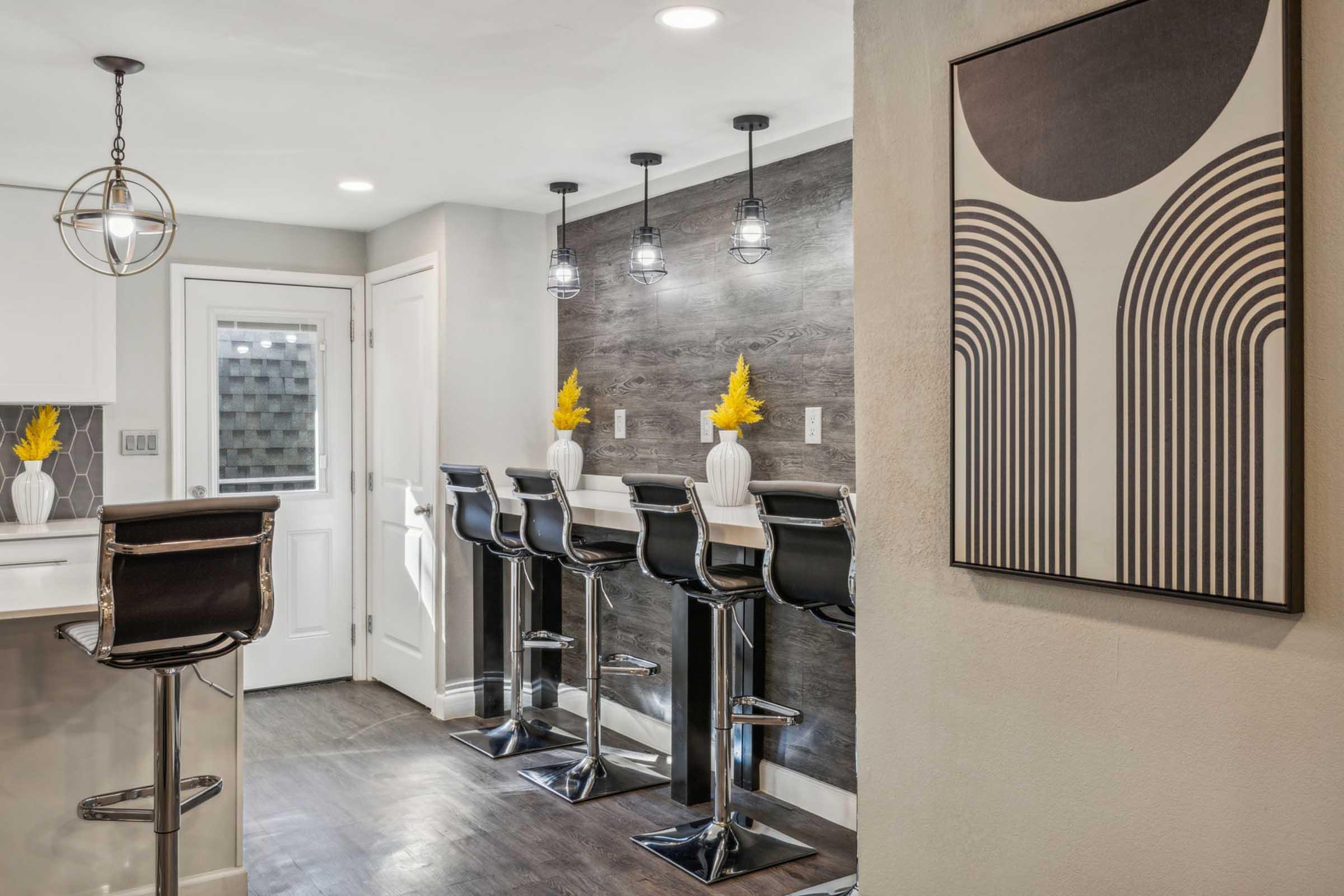 A modern kitchen featuring a bar area with four sleek black stools, a white countertop, and decorative vases with yellow flowers. The background includes a stylish wall art piece and a door leading outside, with warm lighting enhancing the contemporary aesthetic.