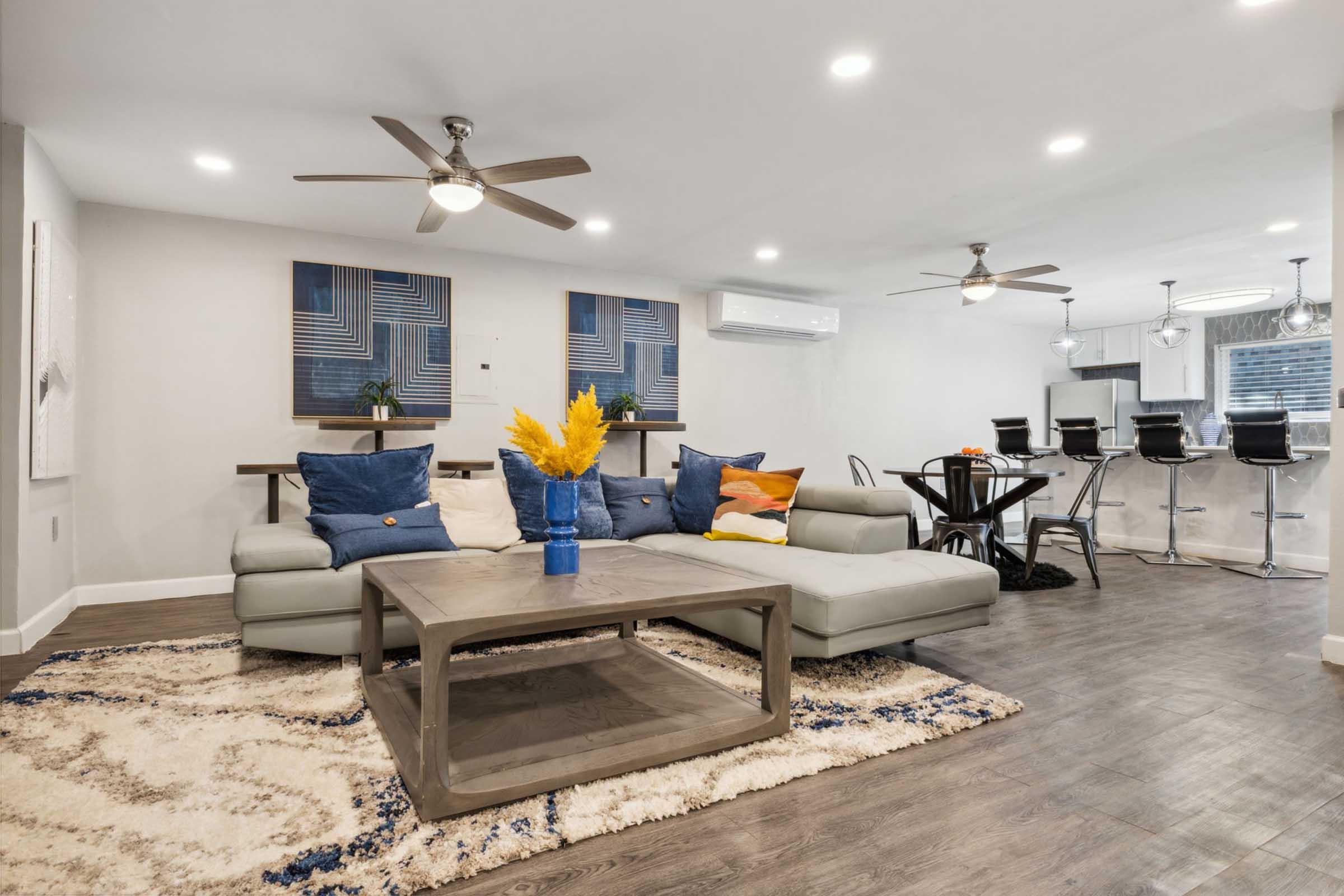 Modern living room featuring a sectional sofa with colorful throw pillows, a coffee table, and a decorative vase with yellow flowers. Two ceiling fans and recessed lighting brighten the space. In the background, there's a dining area with bar stools and contemporary artwork on the walls.