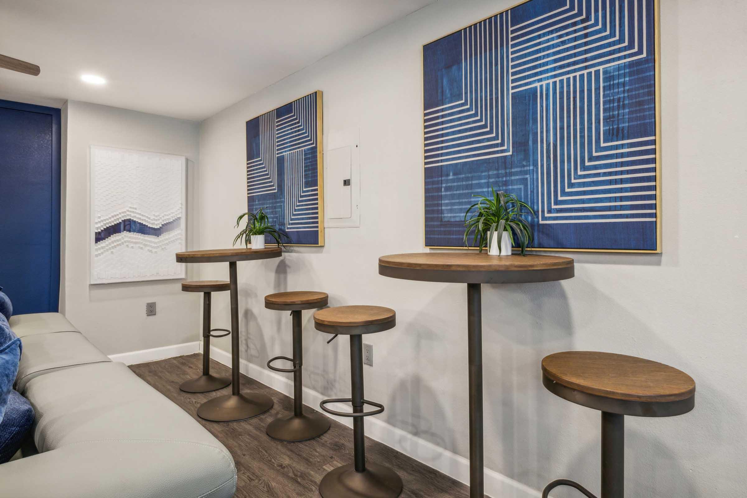 A modern interior featuring a light gray sofa and two abstract blue and white wall art pieces. Four round bar stools with wooden tops and metal bases are positioned near the wall art. The room has a clean, contemporary look with subtle decorations, including small plants on the tables.