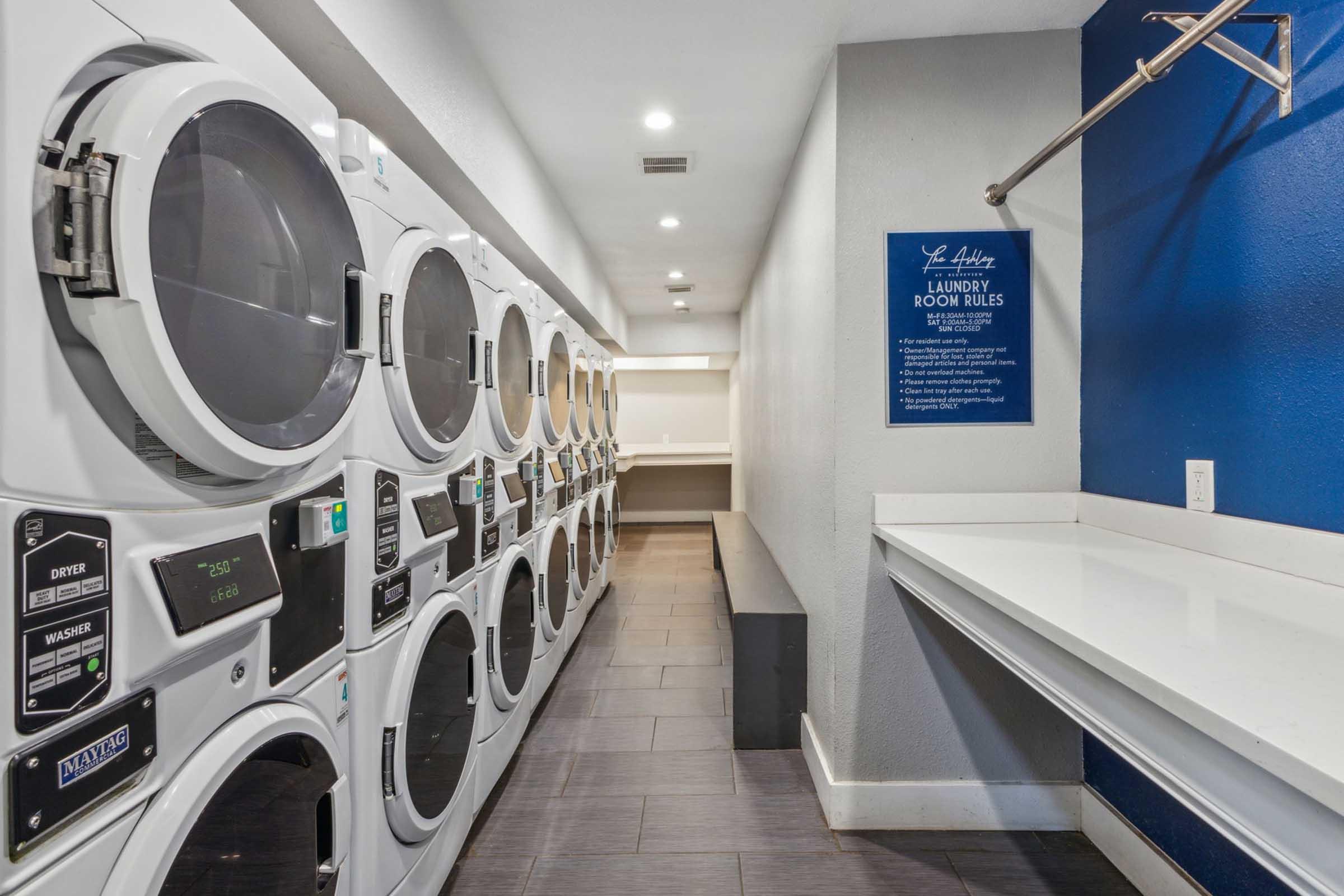 A modern laundry room featuring multiple stacked washing machines and dryers along one wall. On the opposite side, there is a folding counter with a blue wall displaying laundry room rules. The space is well-lit and clean, with tiled flooring and a minimalistic design.