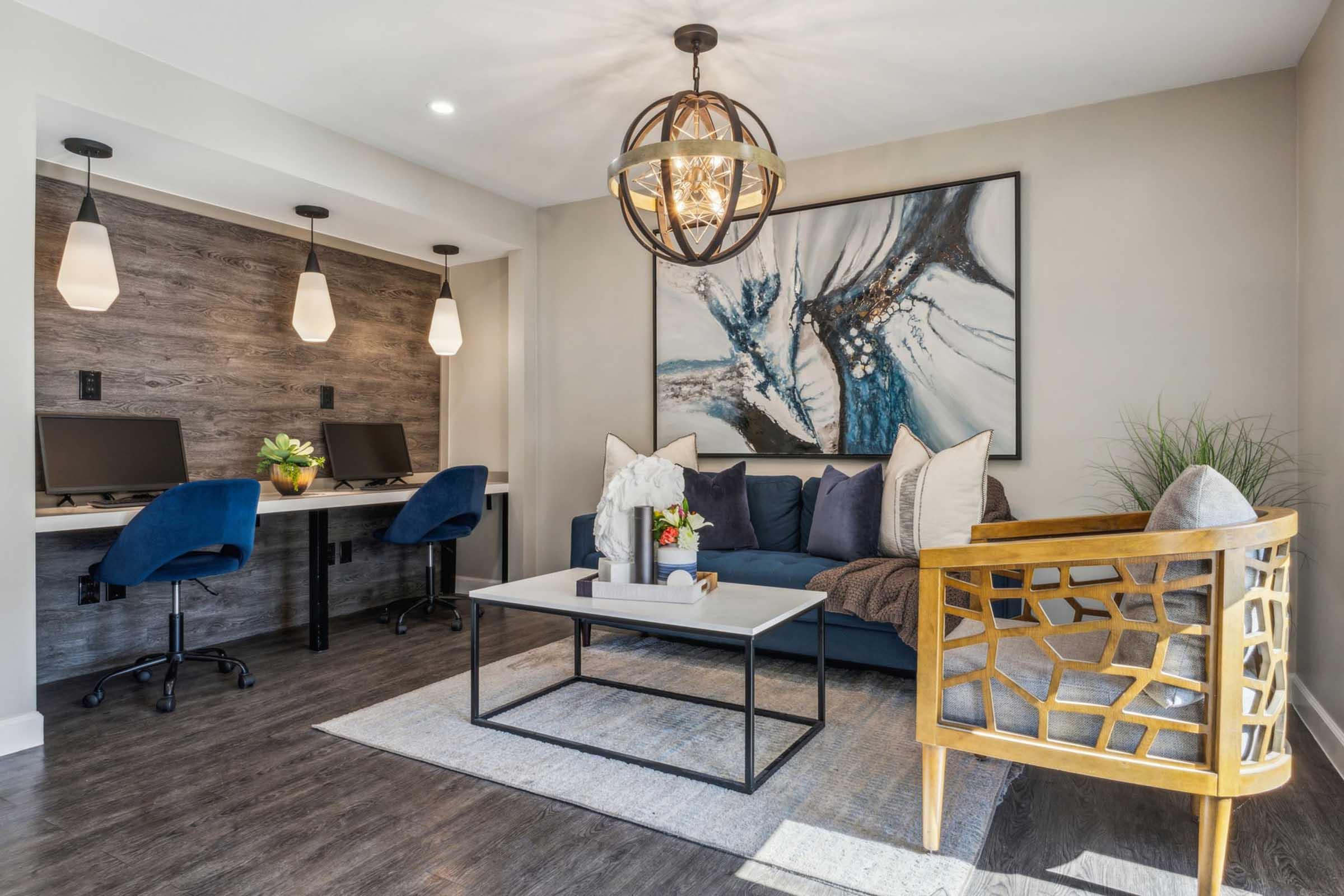 A modern living space featuring a navy blue sofa with decorative pillows, a coffee table on a light rug, and a gold accent chair. The area includes two computer workstations with blue chairs, pendant lighting, and a large abstract artwork on the wall. A plant adds a touch of greenery to the decor.
