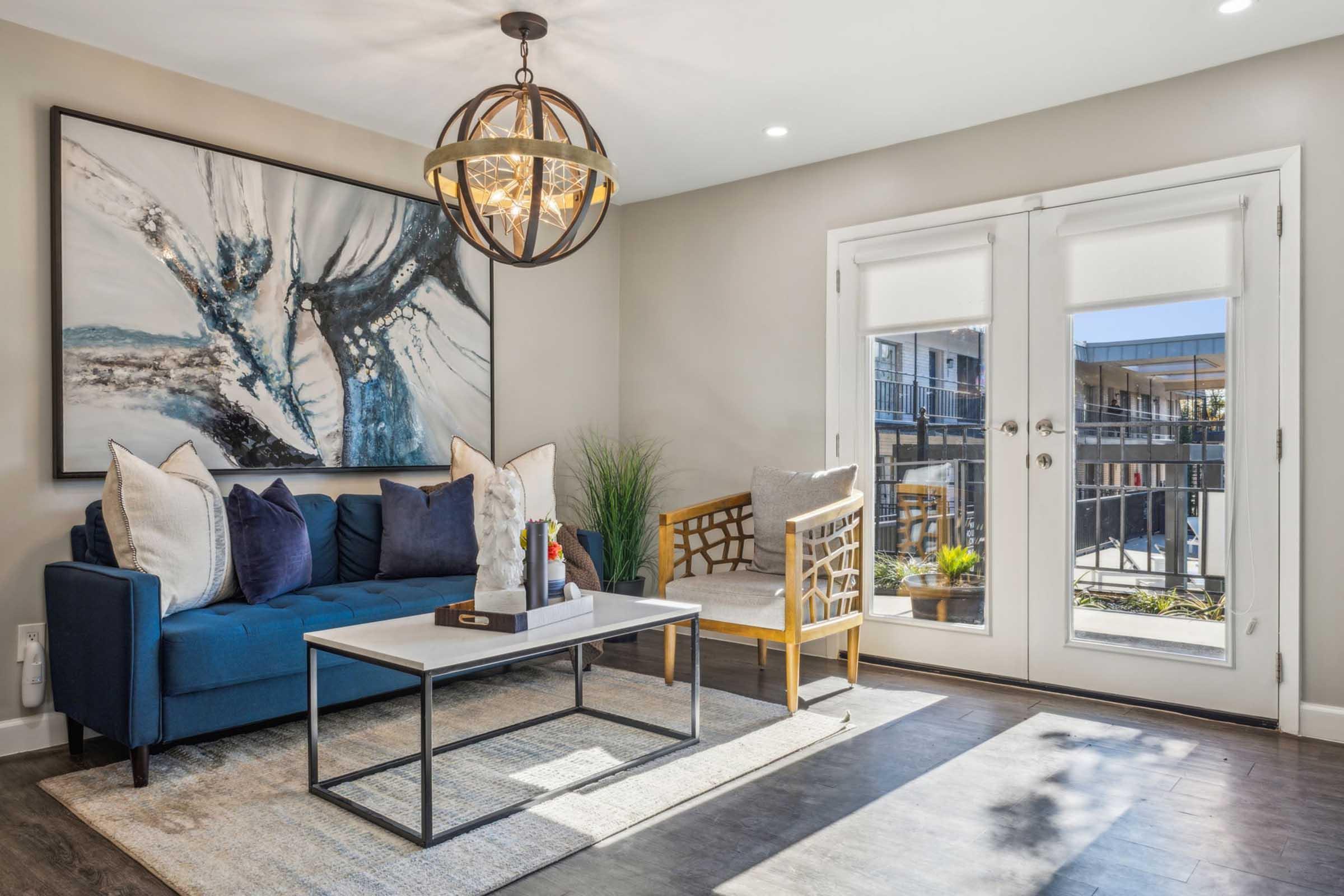 A modern living room featuring a blue sofa with decorative pillows, a stylish coffee table, and a light-colored accent chair. In the background, large doors lead to an outdoor area, while a piece of abstract art hangs on the wall above the sofa, complementing the room's contemporary design.