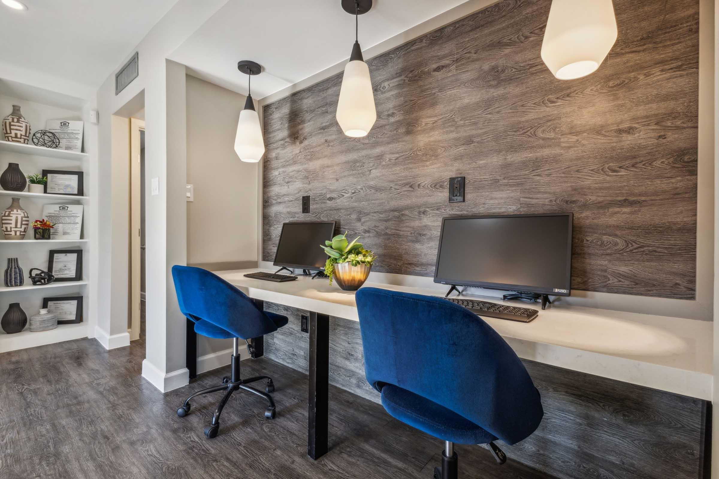 A modern workspace featuring two black desktop computers on a sleek white desk. There are two blue upholstered chairs, and a decorative plant in a gold pot is placed between the computers. The walls are adorned with wood paneling, and there are stylish light fixtures hanging above.