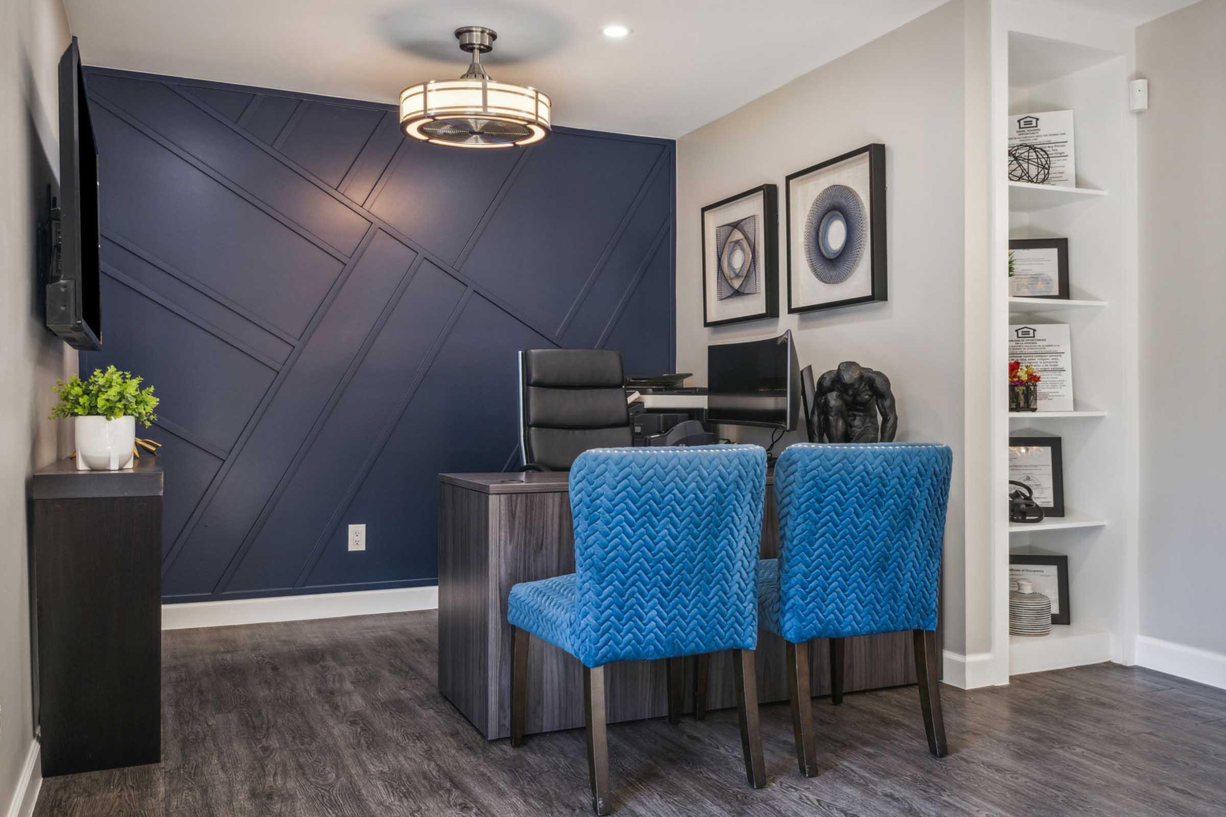 A modern office space featuring a dark blue accent wall with geometric designs. There are two blue upholstered chairs facing a sleek desk, which holds a computer and decorative items. A stylish light fixture hangs overhead, and framed art adorns the walls. Shelves with displayed documents are visible in the corner.