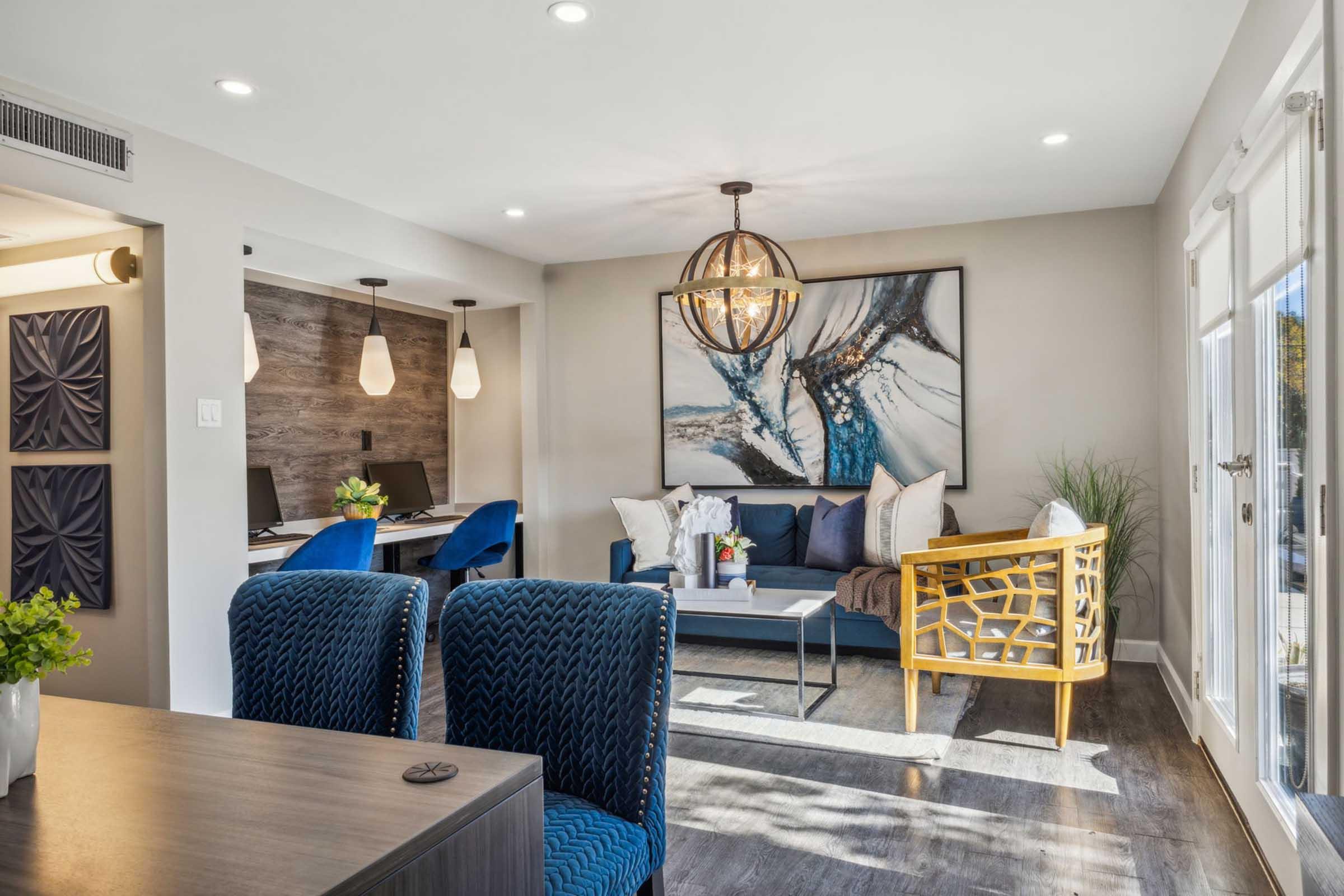 A modern living room featuring a stylish blue sofa with decorative cushions, a gold accent chair, and a wooden coffee table. The space includes a wall art piece with blue and white tones, a desk area with blue chairs, and large windows allowing natural light to brighten the room.