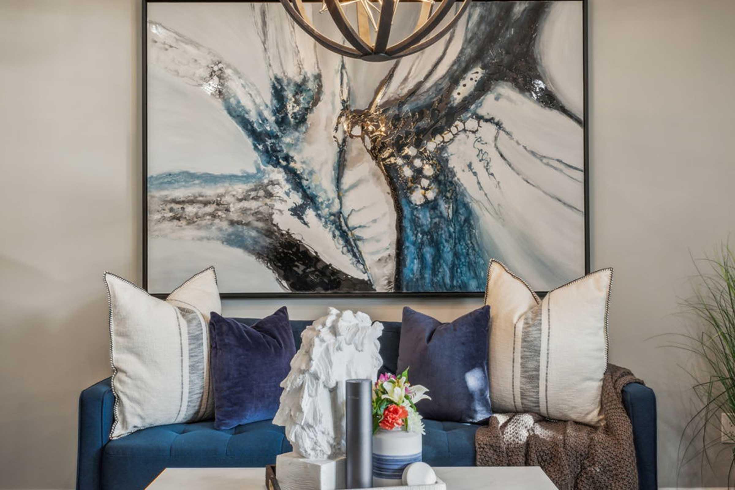 A stylish living room featuring a blue sofa adorned with an assortment of decorative pillows. A modern art piece hangs on the wall, showcasing abstract designs in shades of blue and white. A decorative table displays a floral arrangement and simple decor items, enhancing the contemporary atmosphere.