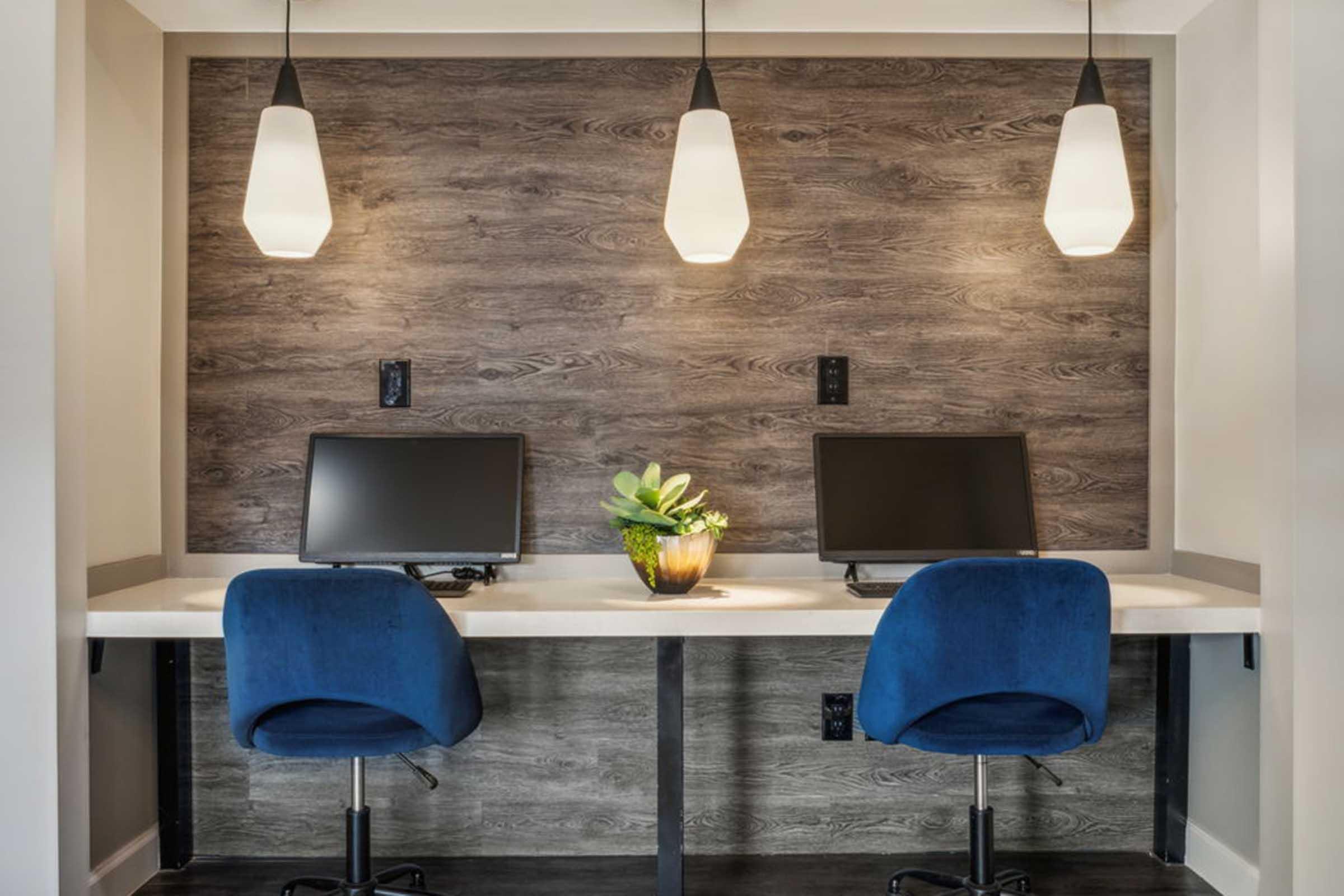 A modern workspace featuring two desktop computers on a white countertop, with blue office chairs. The backdrop consists of textured wooden panels, and three pendant lights hang above, illuminating a small potted plant on the desk. The overall design is sleek and minimalistic.