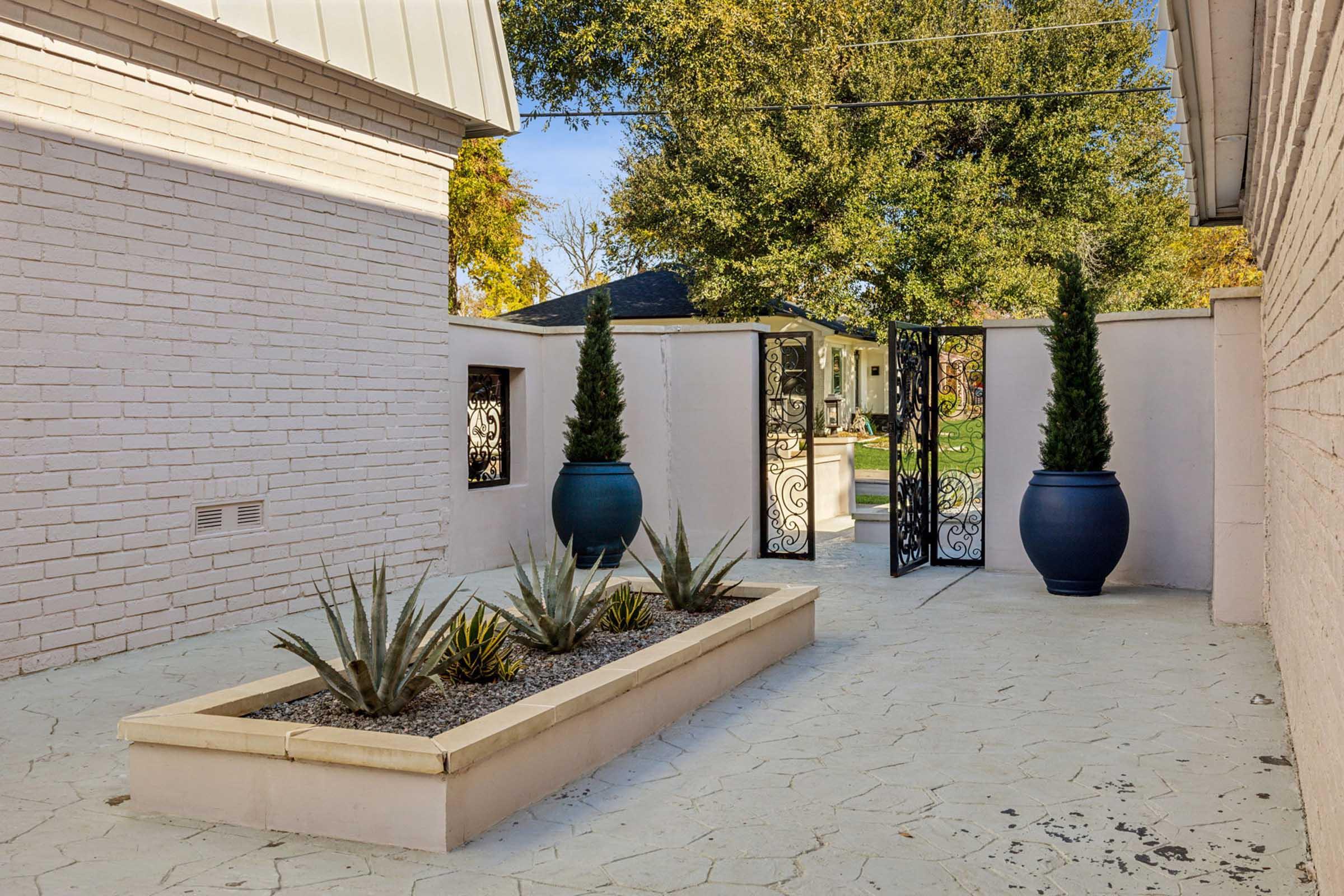 A small outdoor courtyard featuring a raised planter with desert plants, two tall potted trees flanking ornate black iron gates, and a textured pavement. The background includes trees and a glimpse of a house beyond the gates, illuminated by natural sunlight.