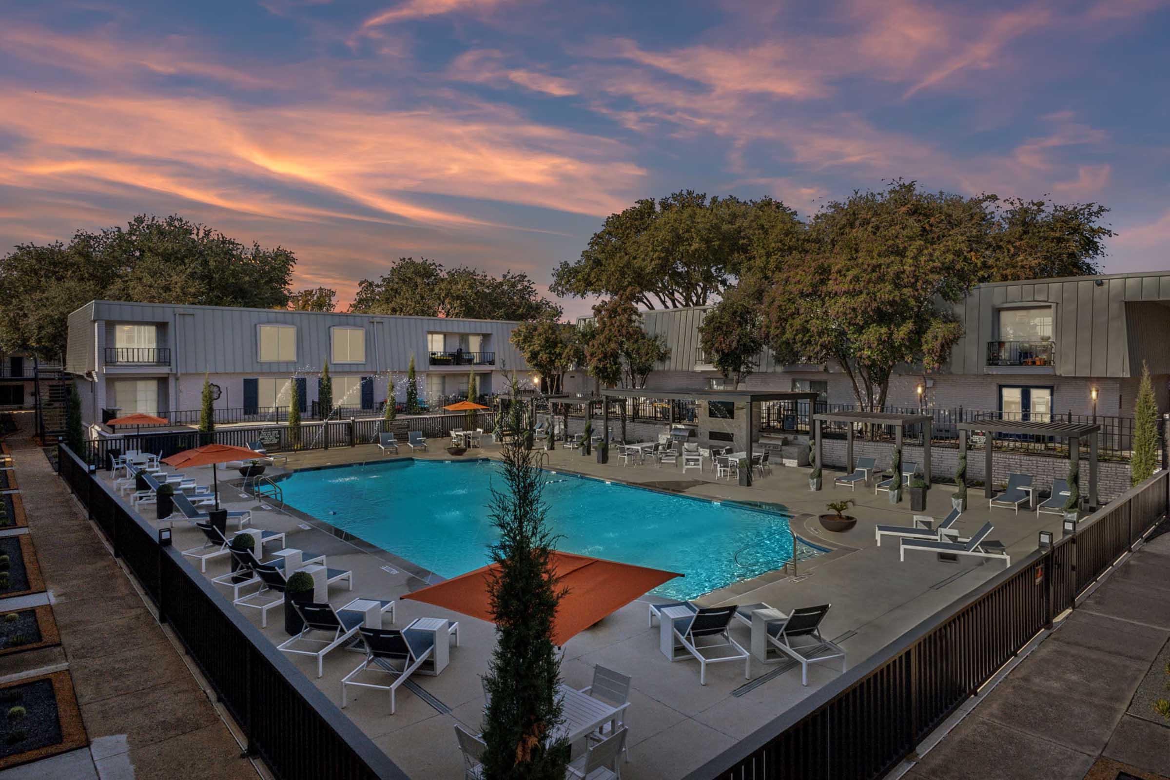 A serene outdoor swimming pool surrounded by lounge chairs, orange umbrellas, and modern buildings. The scene is set against a beautiful sunset sky with vibrant colors, creating a relaxing atmosphere. Lush green trees frame the pool area, adding to the tranquil ambiance.