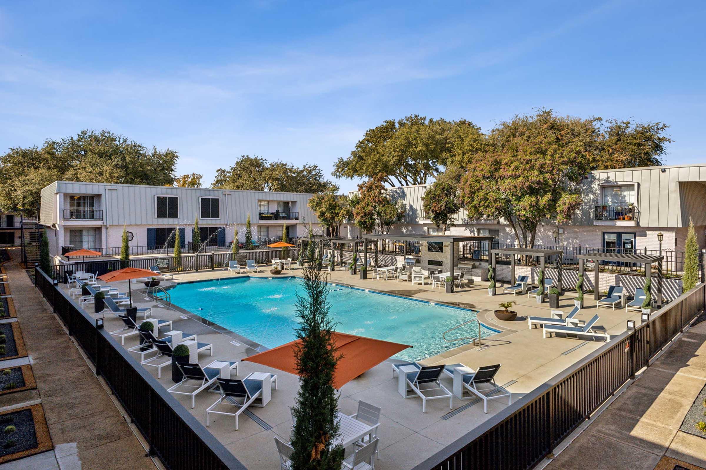 A spacious outdoor pool area surrounded by lounge chairs and orange umbrellas, set within a modern apartment complex. The scene includes trees providing shade, and there are two-story buildings in the background, indicating a welcoming residential environment. Clear blue sky overhead.