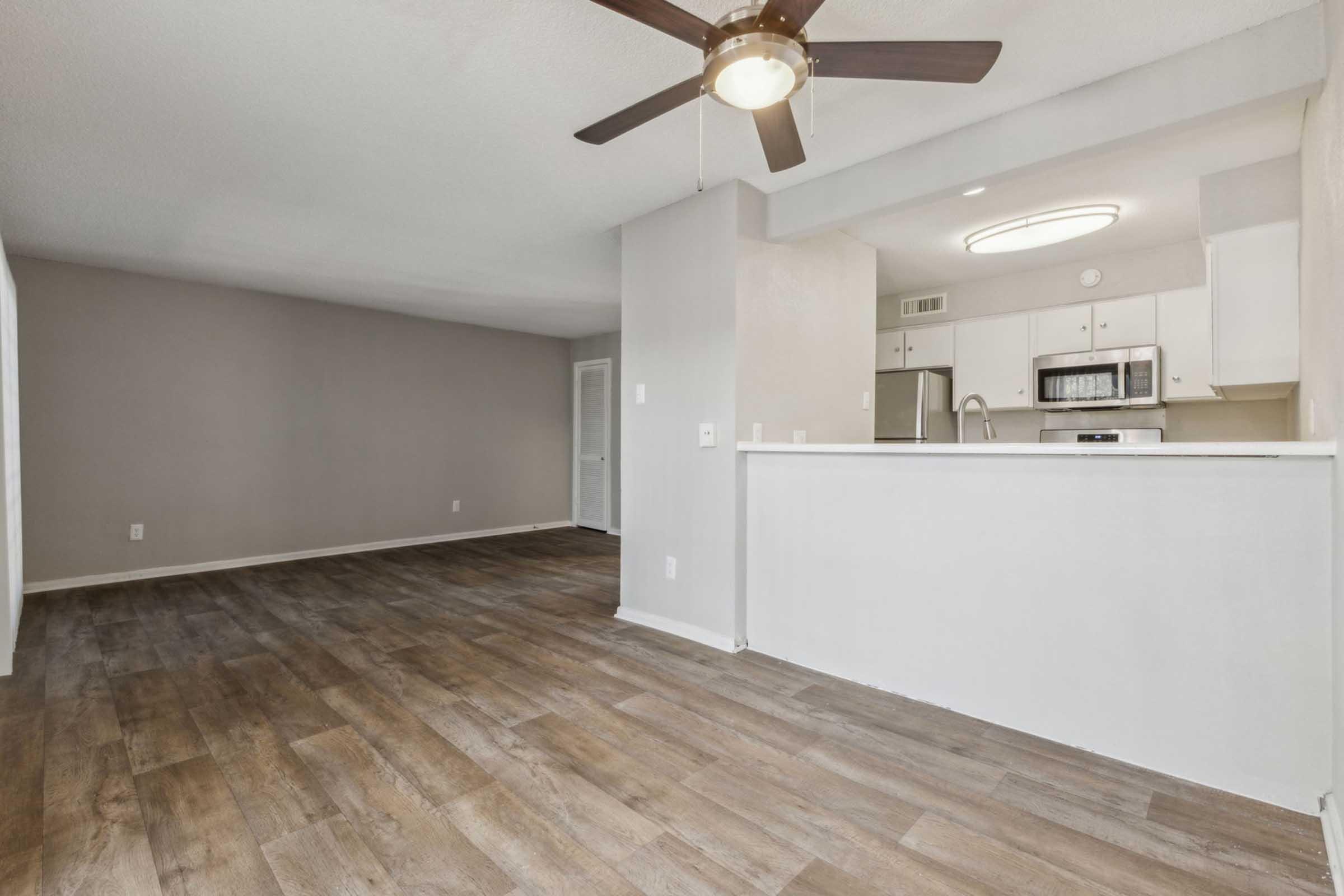 A spacious living area with light-colored walls and wood-like flooring. The open layout features a ceiling fan, leading to a kitchen area with white cabinets, a microwave, and a countertop. Natural light enhances the inviting atmosphere, making the space feel warm and welcoming.