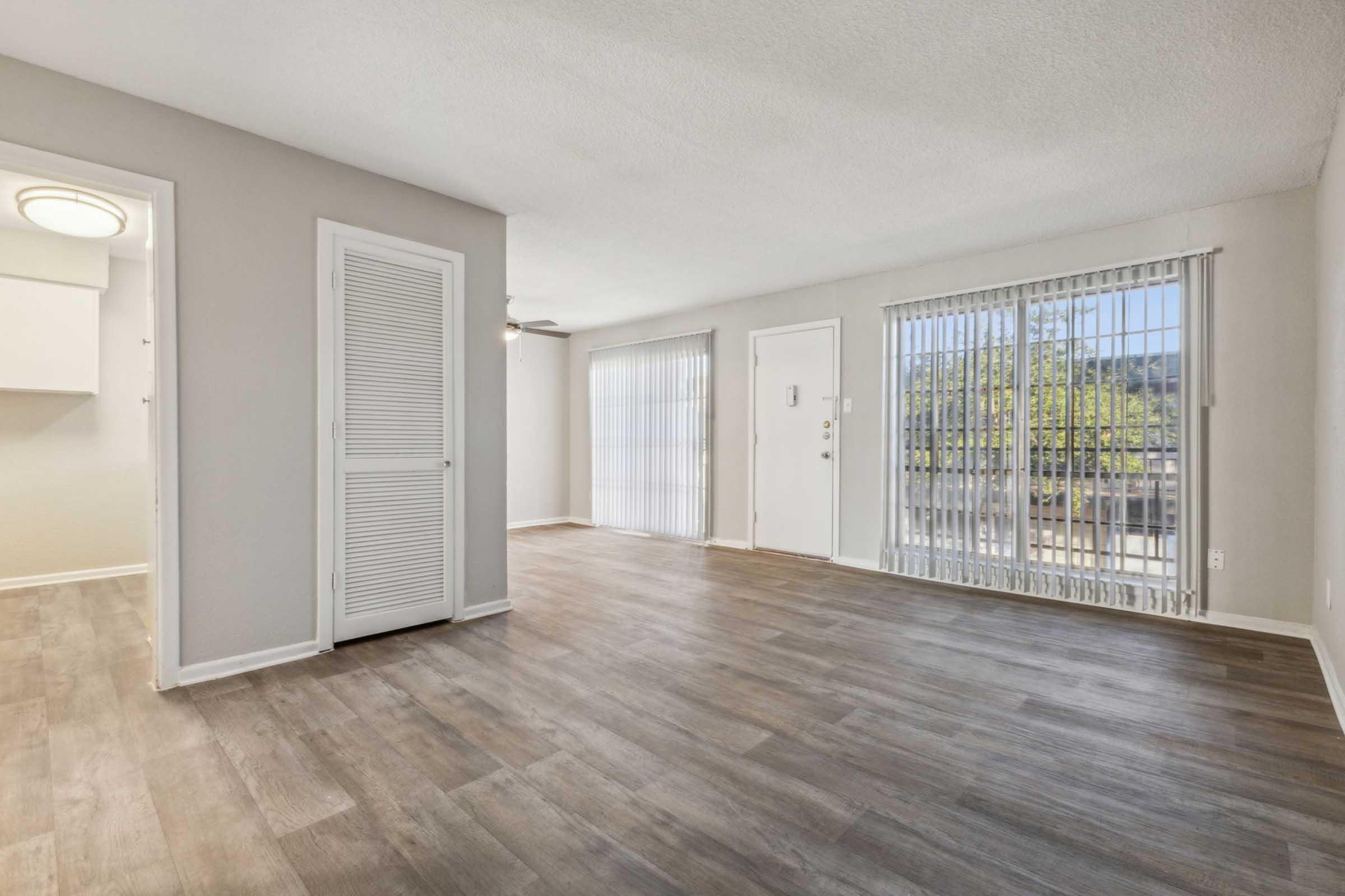 A spacious, well-lit living room with light-colored walls and modern wood-like flooring. Large windows with vertical blinds let in natural light. There's a door leading outside and a closet with louvered doors on the left. A ceiling fan is visible on the left side, adding to the airy ambiance.