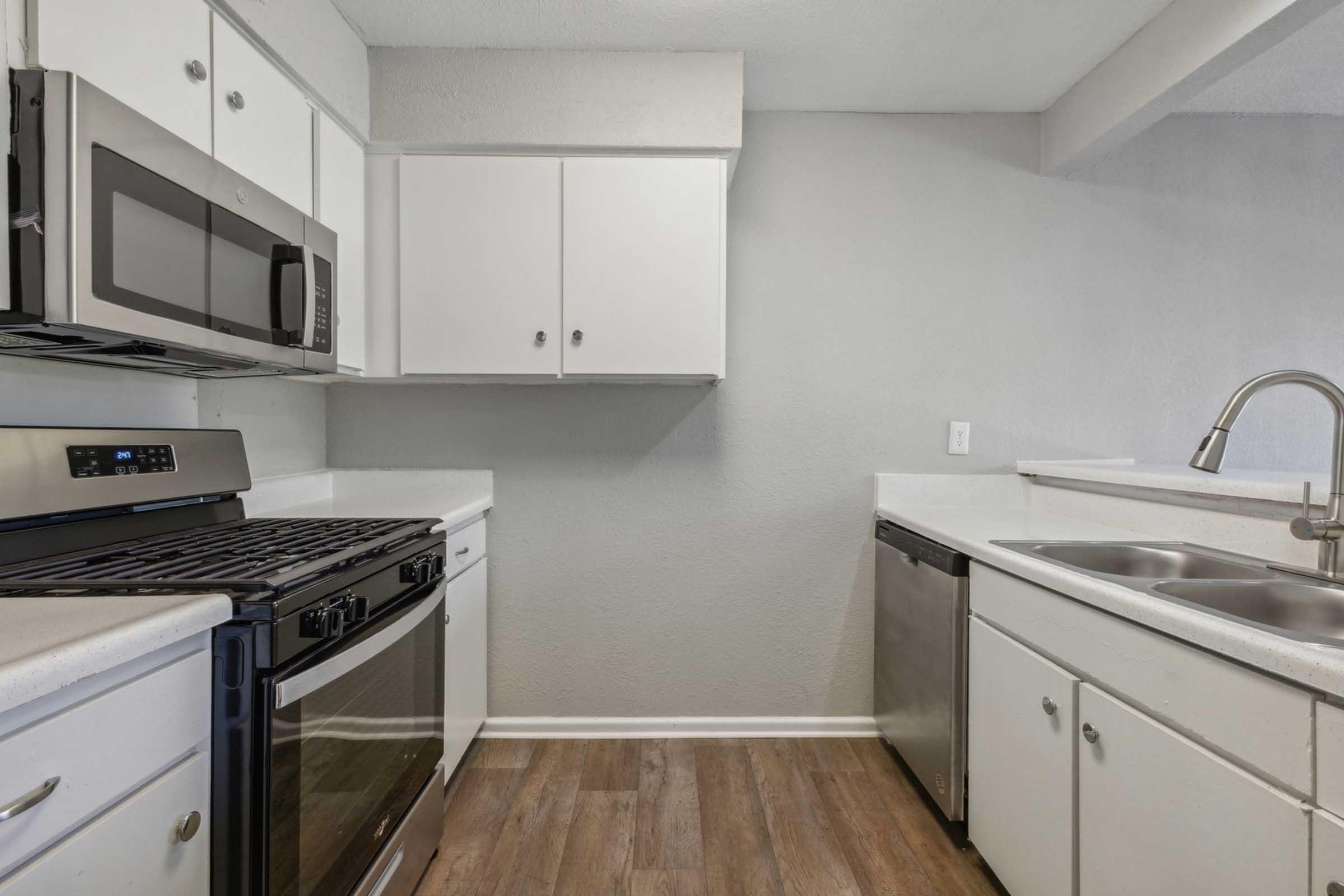 Modern kitchen with white cabinets, stainless steel appliances, a gas stove, and a microwave. It features a double sink with a sleek faucet and laminate flooring. The walls are painted in a light gray color, creating a bright and open atmosphere. There are no items on the countertops, giving a clean and uncluttered look.